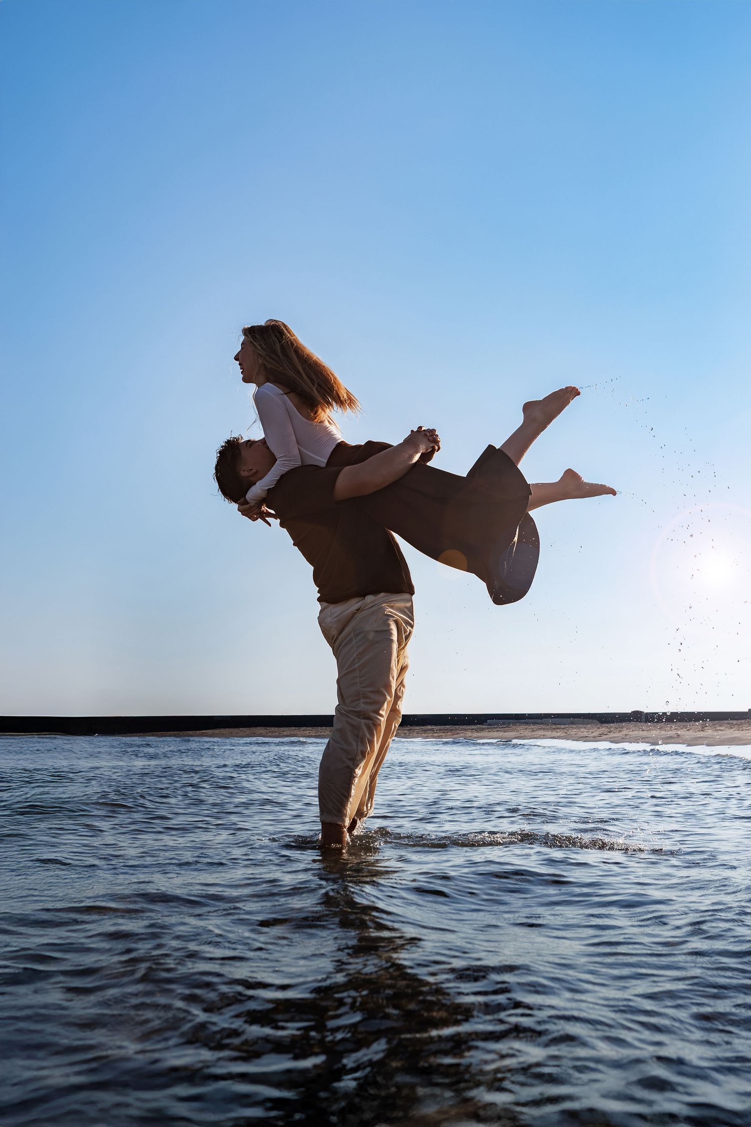 Two people recreating an iconic dance lift pose in shallow ocean water at sunset against a blue sky.