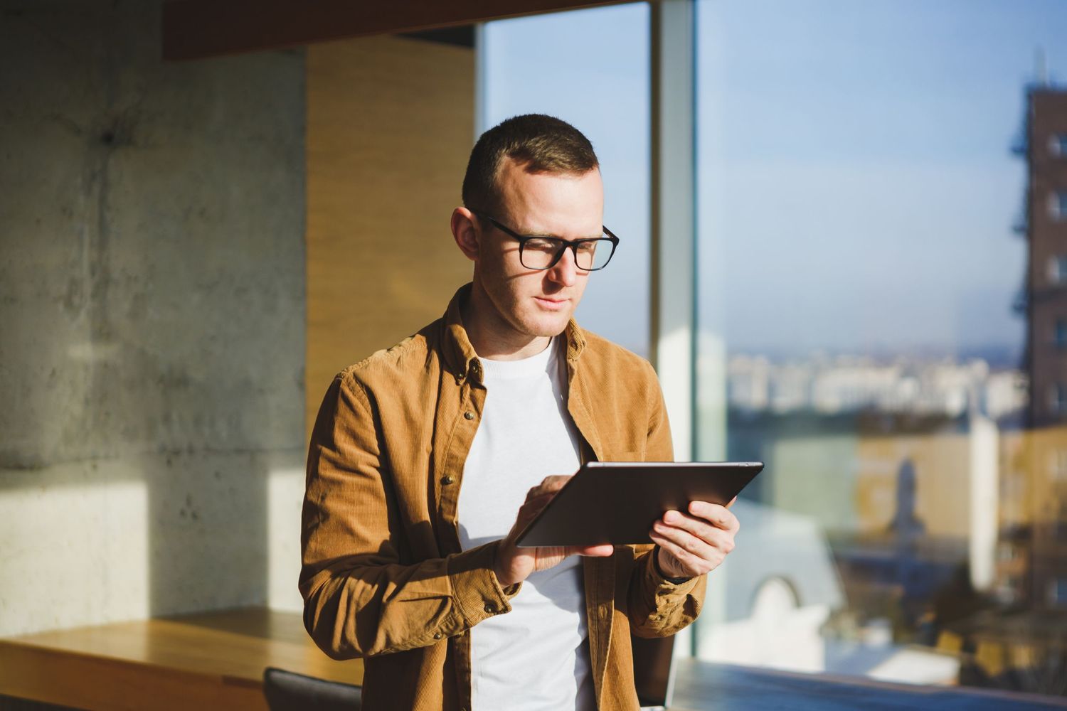 Business person in casual attire working on digital tablet near sunny window.