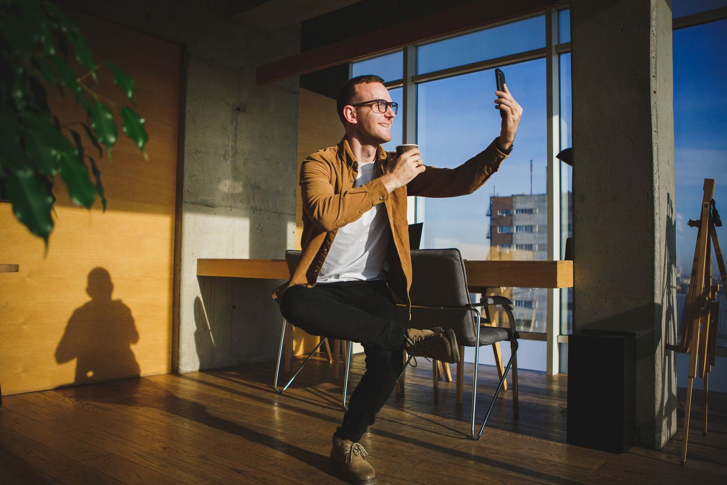 Person in brown jacket using mobile device while standing in sunlit office space.
