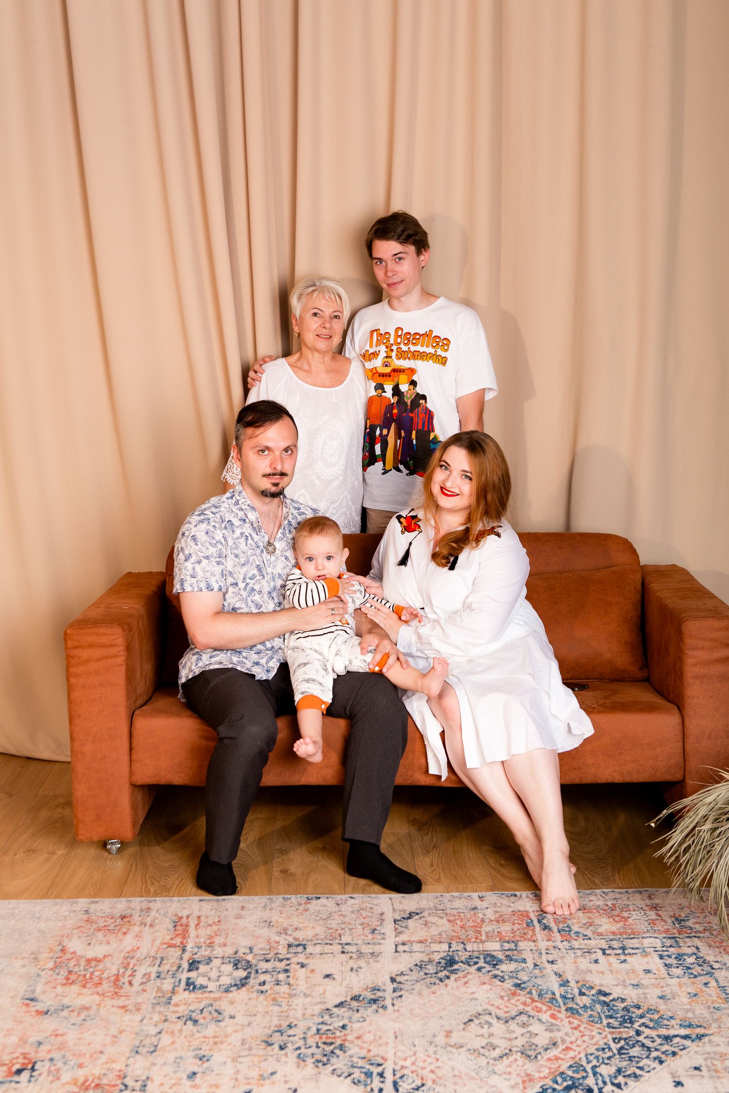 A family gathering on a brown sofa in a living room with decorative curtains and a patterned rug.