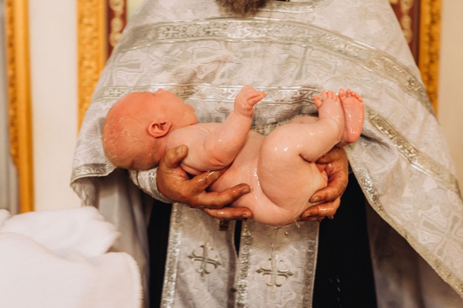 Baby being held in ornate religious robes during Orthodox Christian baptism ceremony.