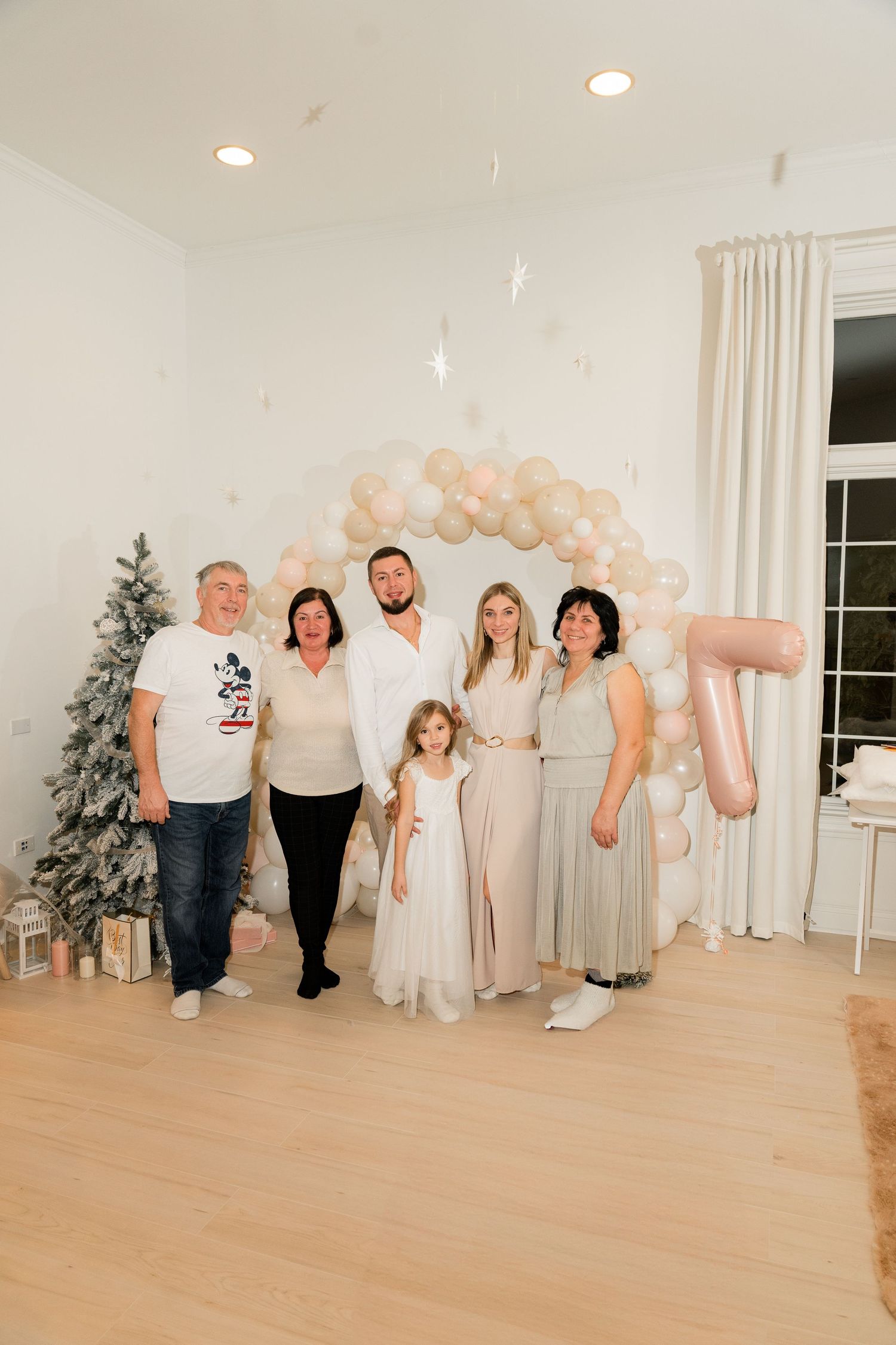 Family gathering under a pink balloon arch with a Christmas tree and presents in the background.