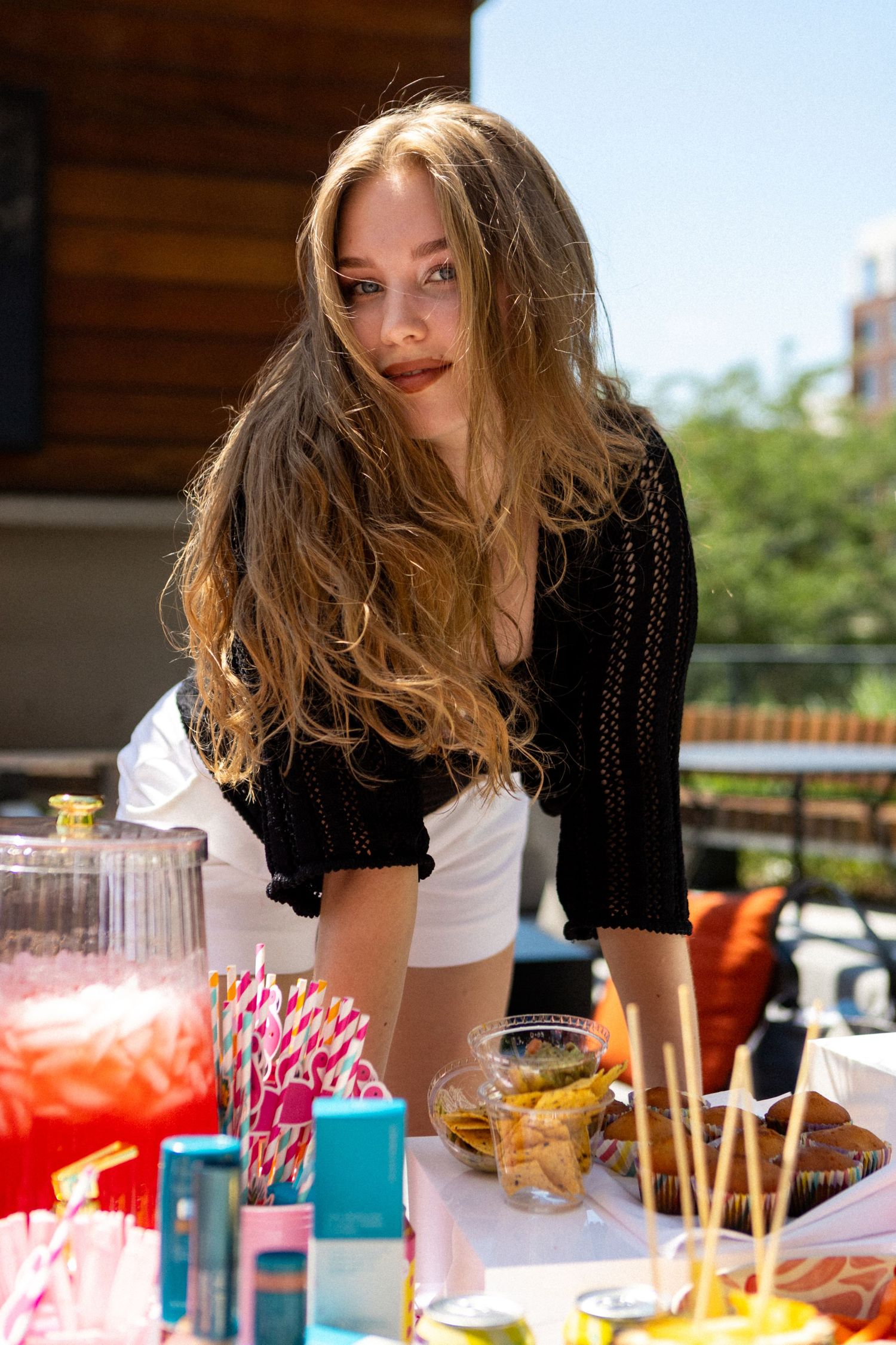 Woman in black and white outfit admires colorful cocktails and appetizers at an outdoor restaurant patio.