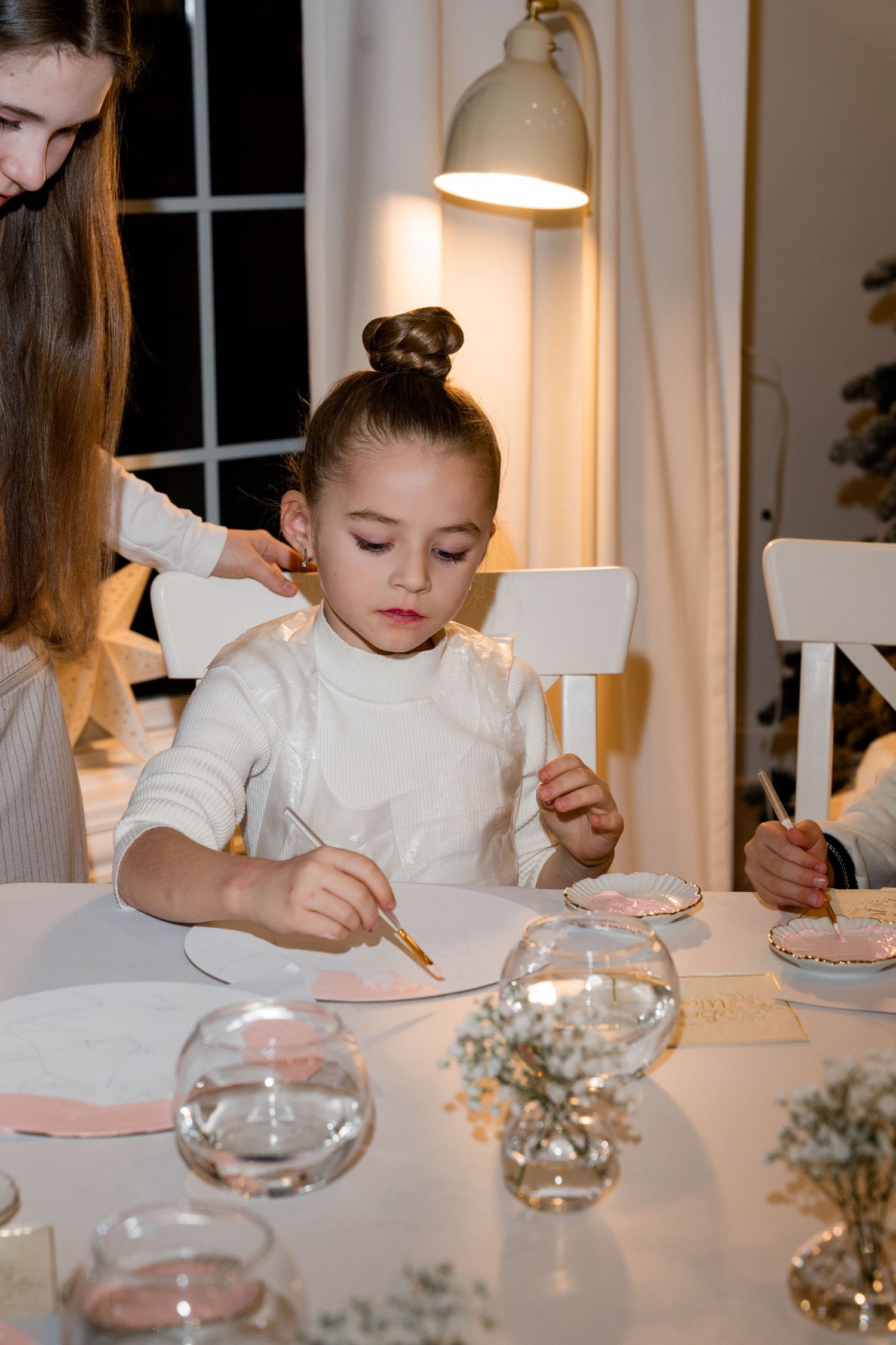 Children doing crafts at white table with festive decorations and soft lighting.