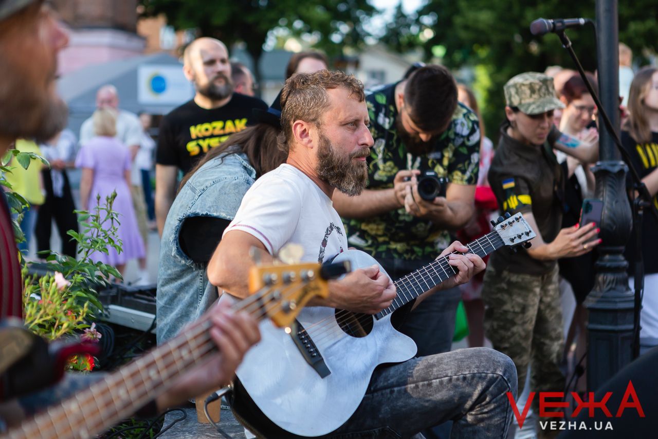 Musicians playing acoustic instruments during an outdoor performance with crowd in background.