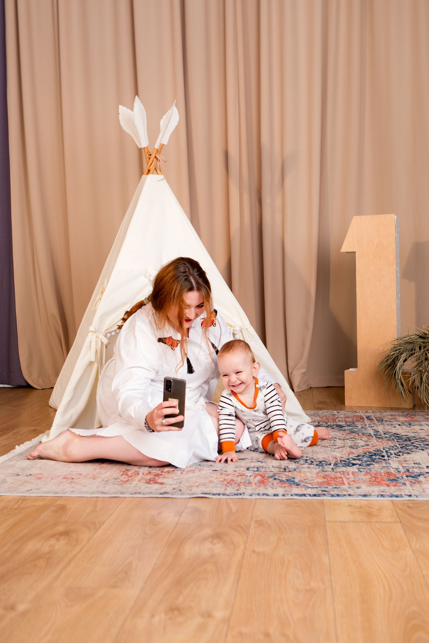 A small child and adult relax on a rug near a teepee tent while looking at a phone.
