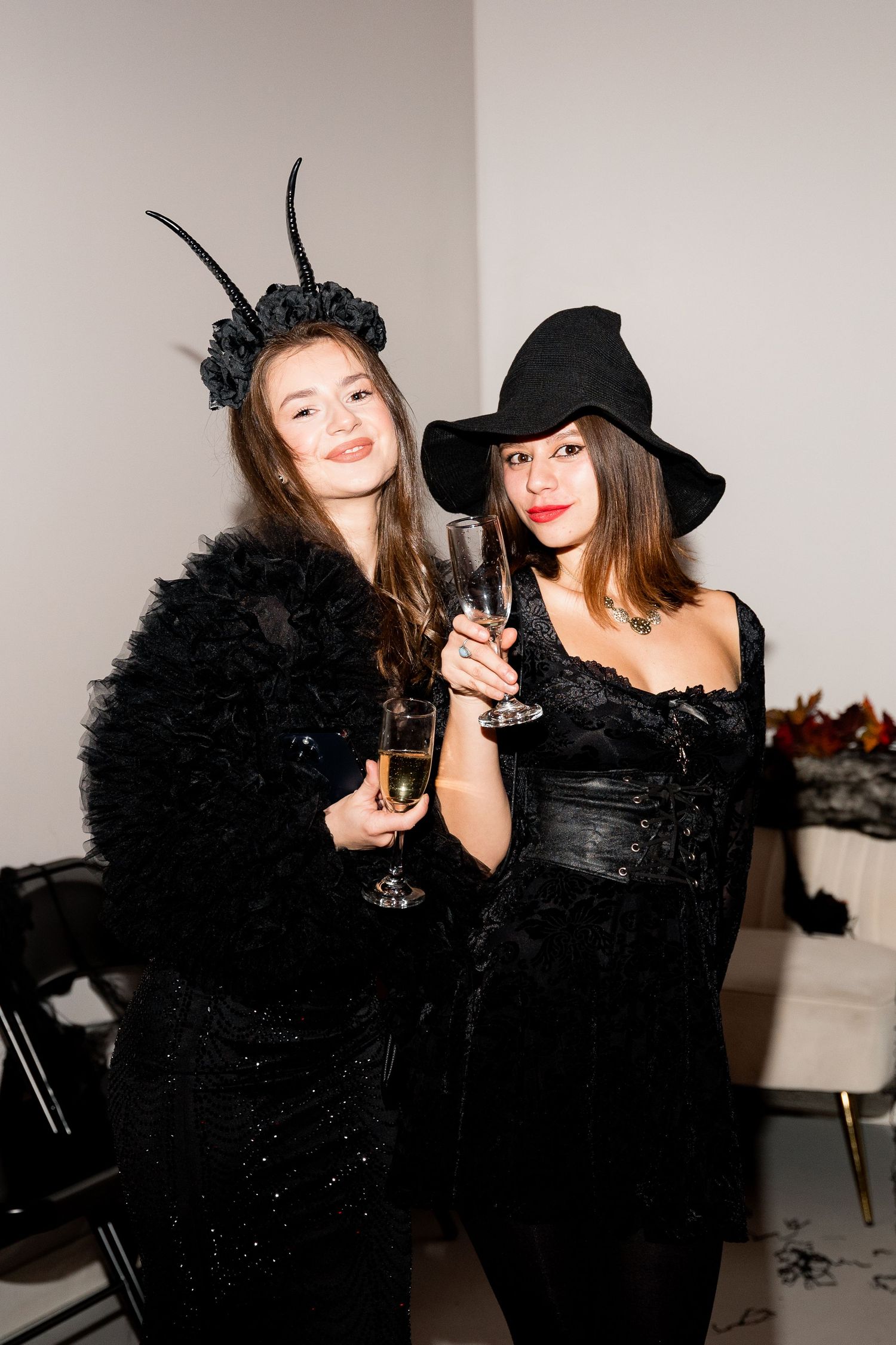 Two people in dark Halloween costumes hold champagne glasses at a party.