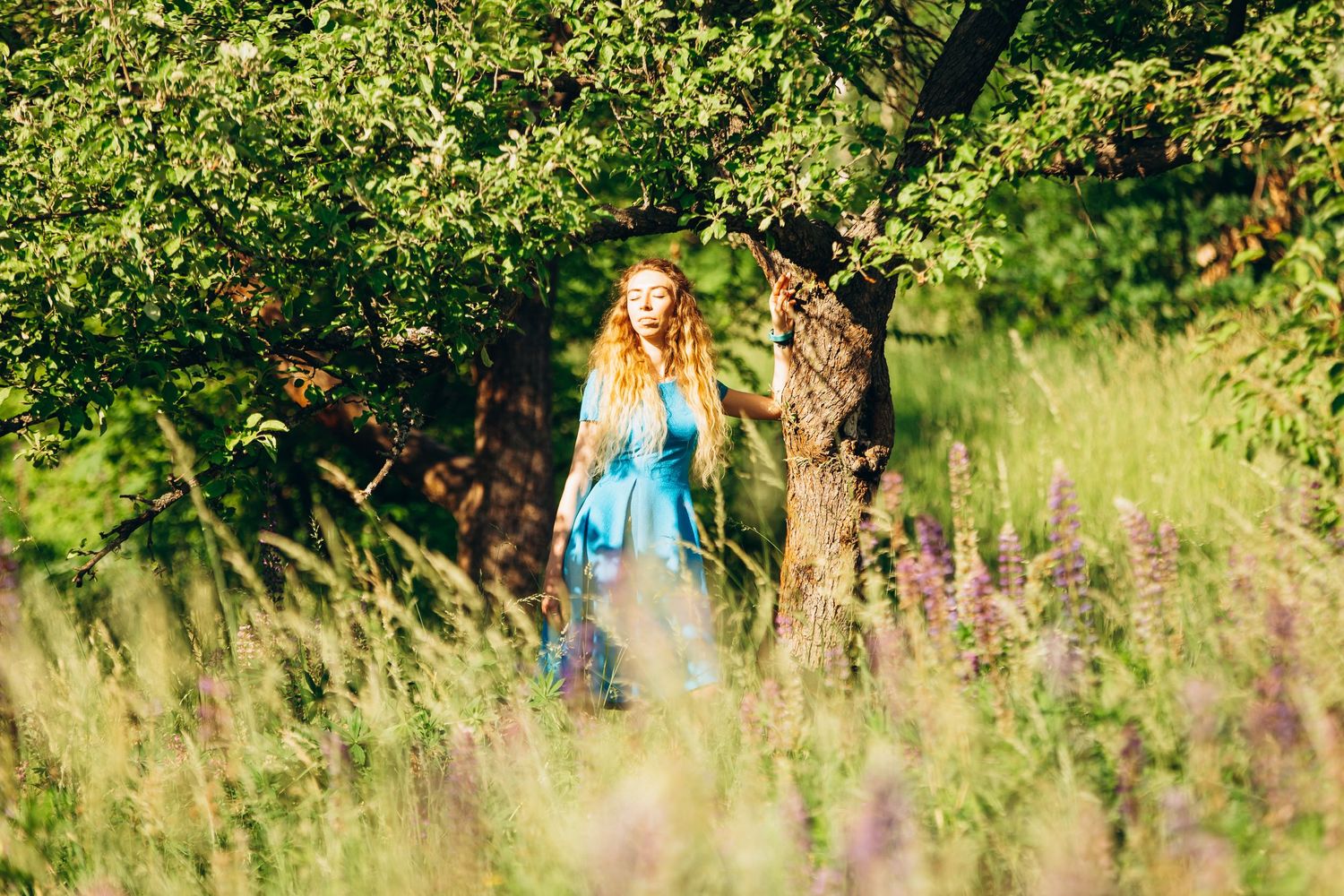 Person in flowing blue dress walking through tall grass and wildflowers in sunlit garden.