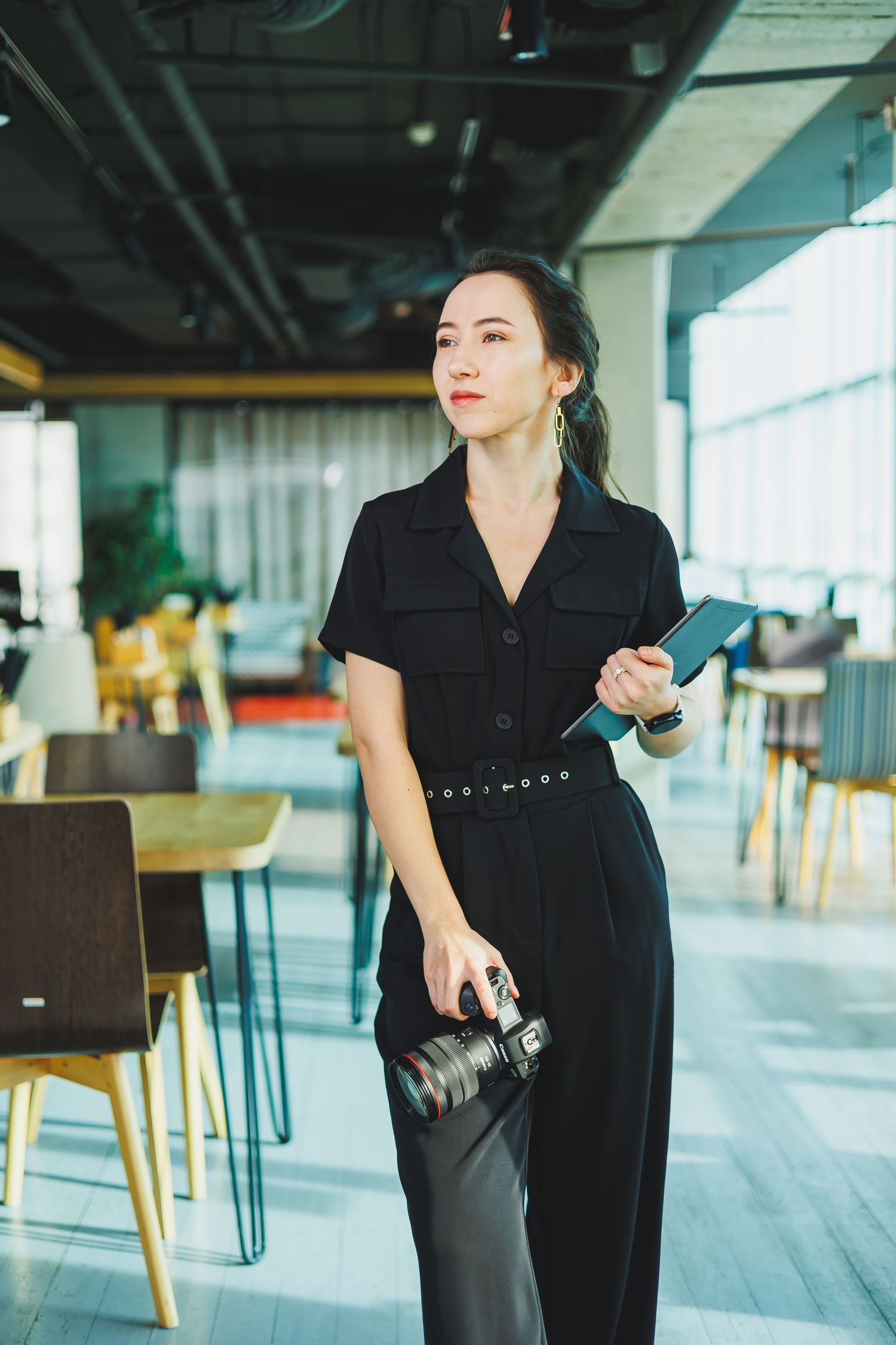 A photographer in a black jumpsuit carries camera gear while standing in a modern office space.