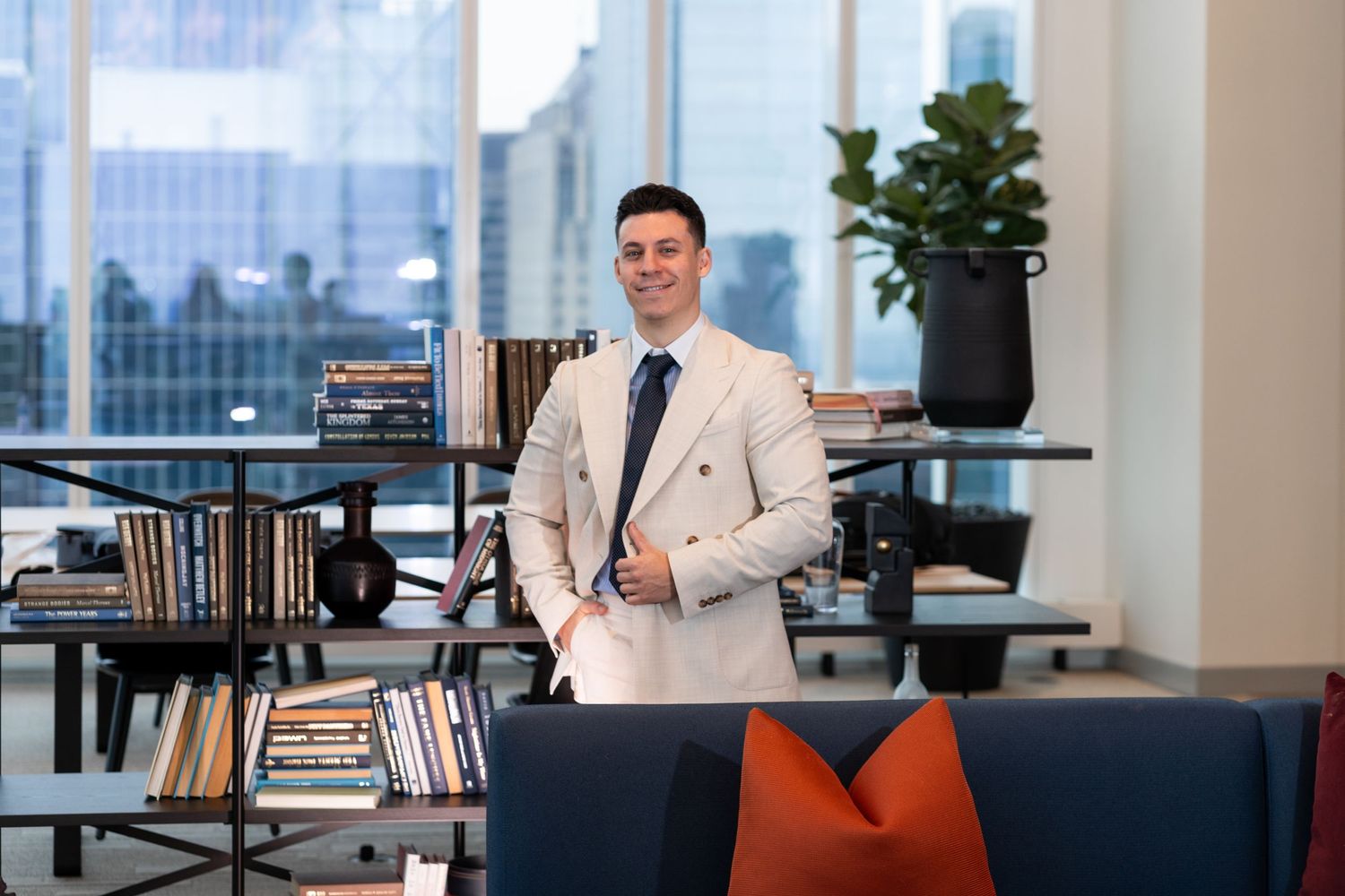Professional in cream suit standing in modern office lounge with city views and bookshelves.