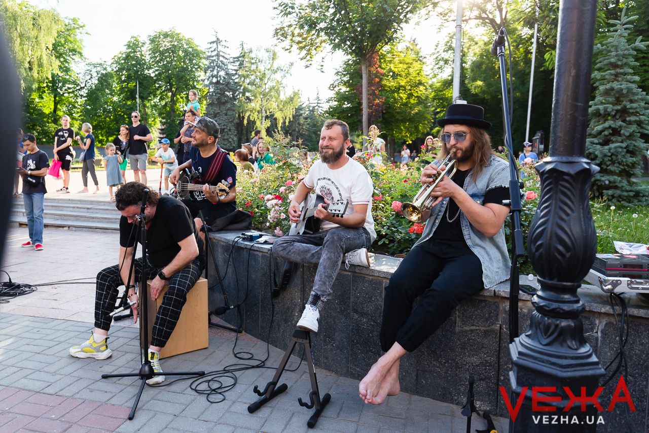 Musicians perform outdoors on benches in a park setting during summer.