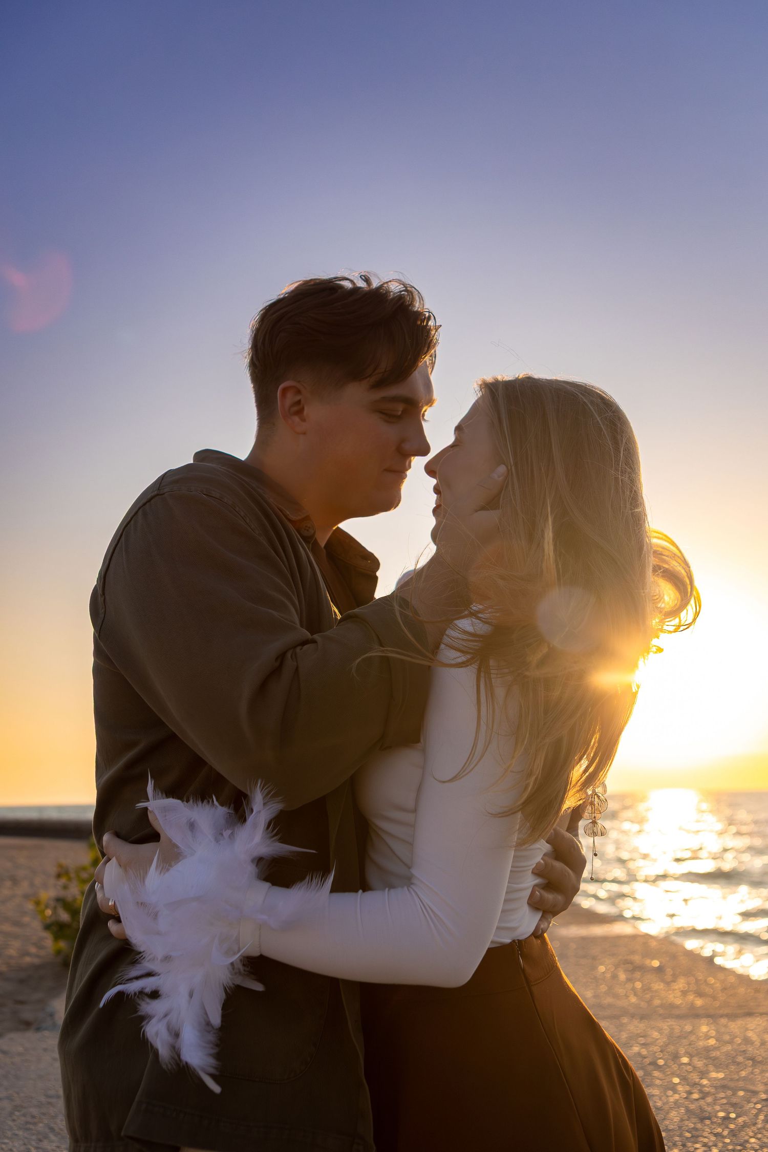 A couple embraces on a beach at sunset with ocean waves and golden light creating a romantic silhouette.