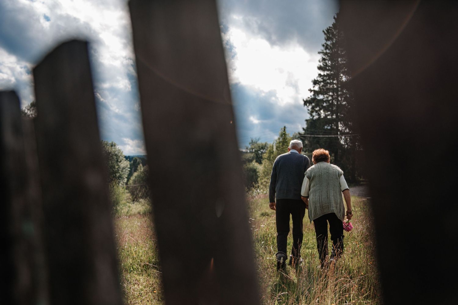 View through fence posts of two people walking together in a sunny meadow.