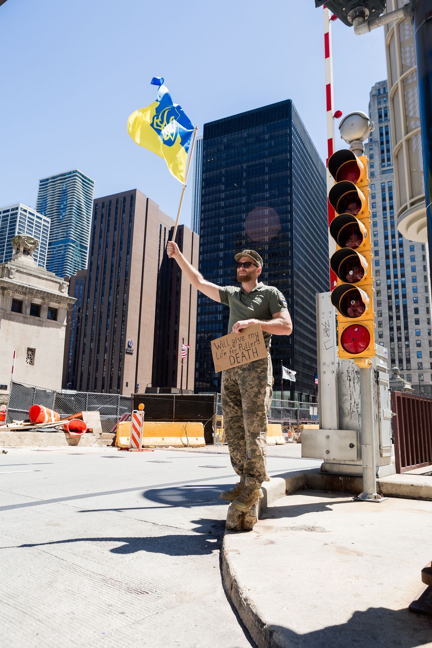 Protester waves flag in urban city setting next to traffic light pole with tall buildings and blue sky in background.