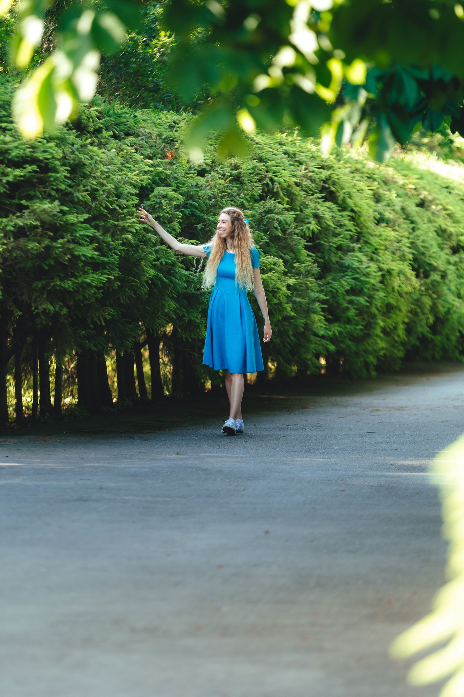 Person in blue dress walking along tree-lined path in dappled sunlight.