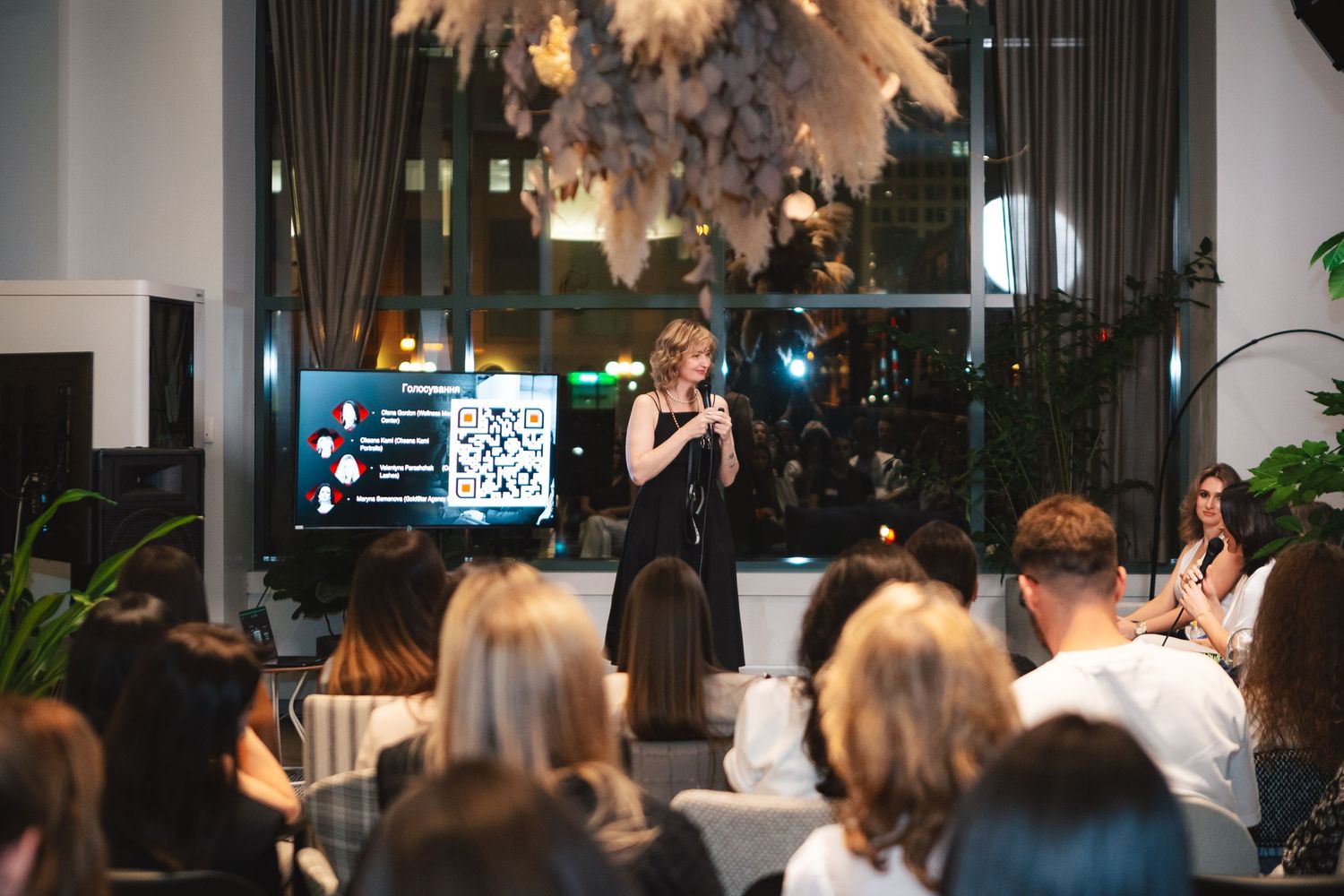 Speaker presenting to an engaged audience under decorative pampas grass hanging installation at modern venue.