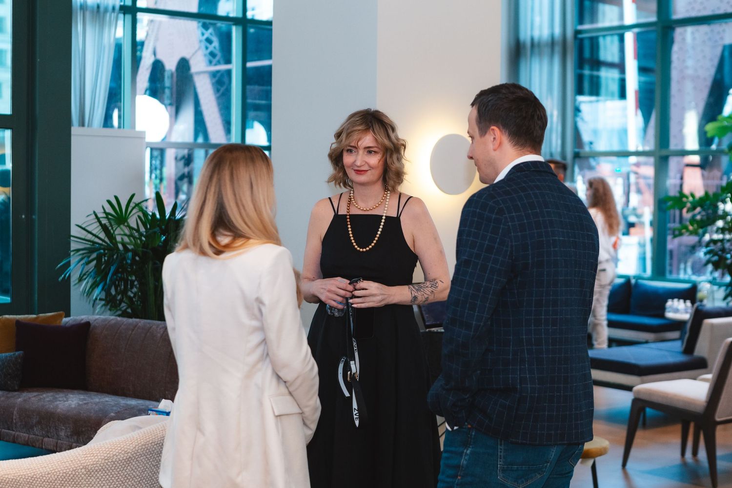 Business professionals engaged in conversation in a modern office lobby with blue-tinted windows and plants.
