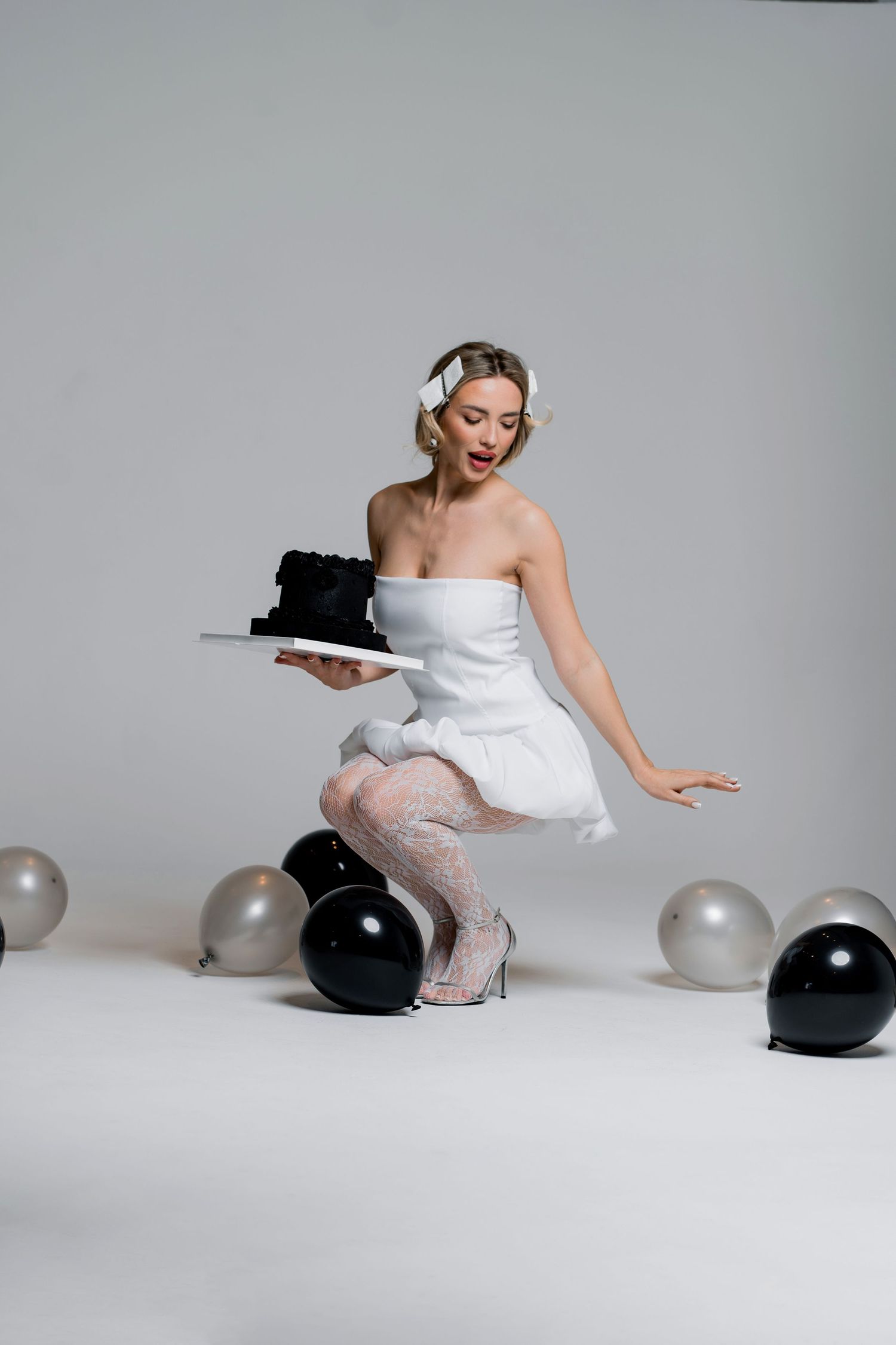 A lady in white dress crouches among black and silver balloons in a studio setting.