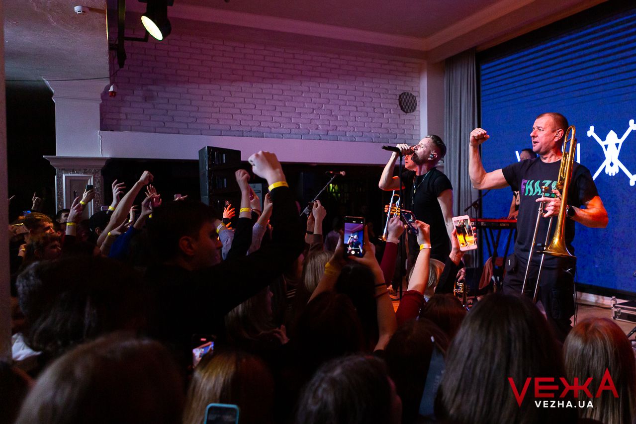 Enthusiastic concert crowd raises hands while filming performers on stage with phones.