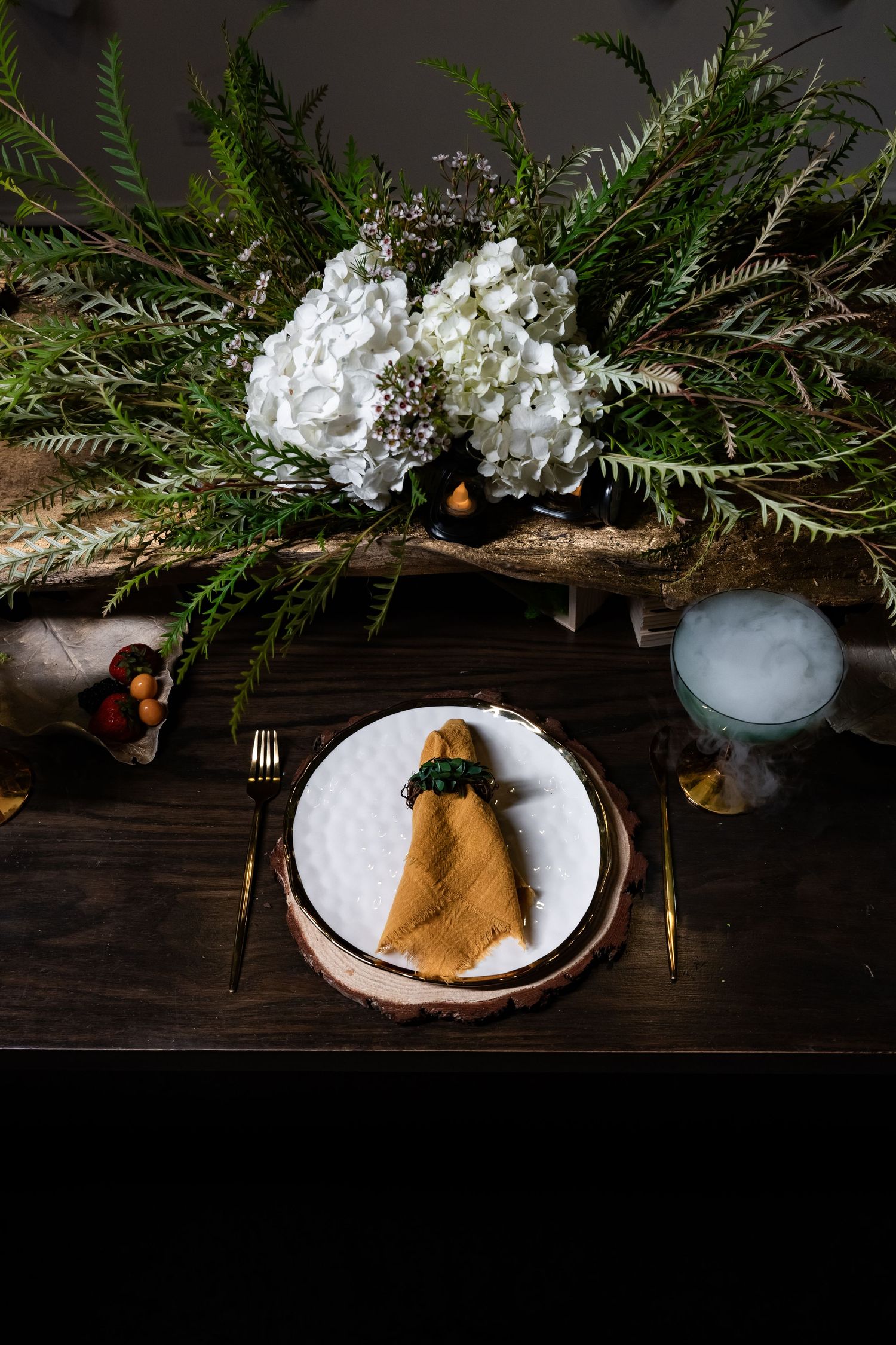 Elegant table setting with fern decor, white hydrangeas, and gold-accented place setting on dark wooden surface.
