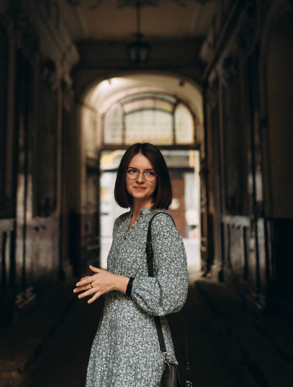 Person in floral dress and glasses standing in historic archway with vintage architecture.