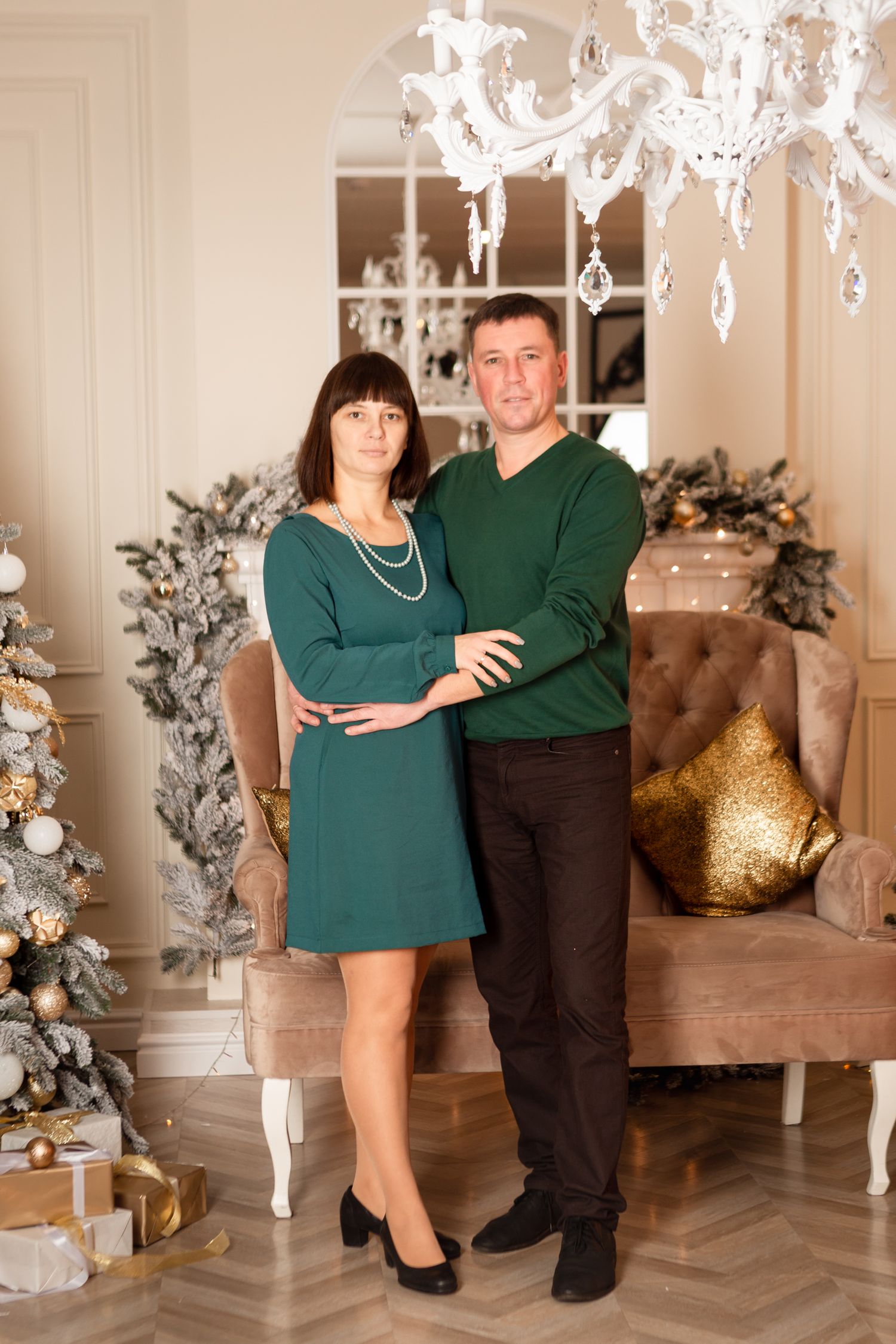Couple in matching green outfits poses by decorated Christmas tree and elegant chandelier.