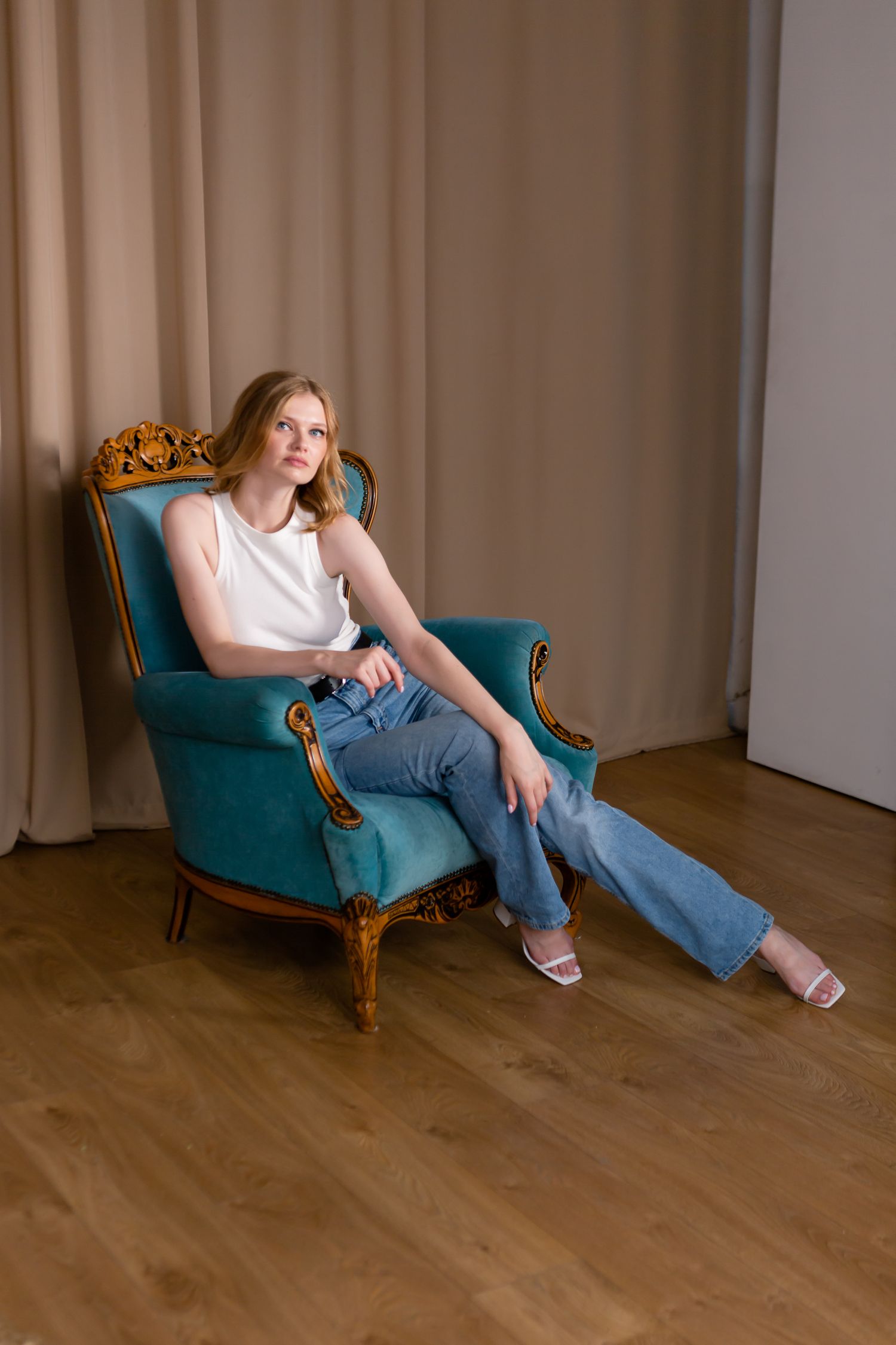 Person relaxing in a vintage teal armchair wearing casual white top and jeans in an interior setting.