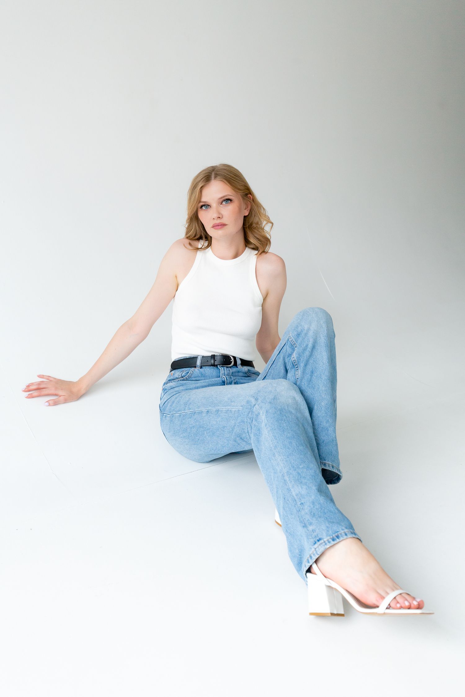 Model in white tank top and light blue jeans poses against white background with white sandals.