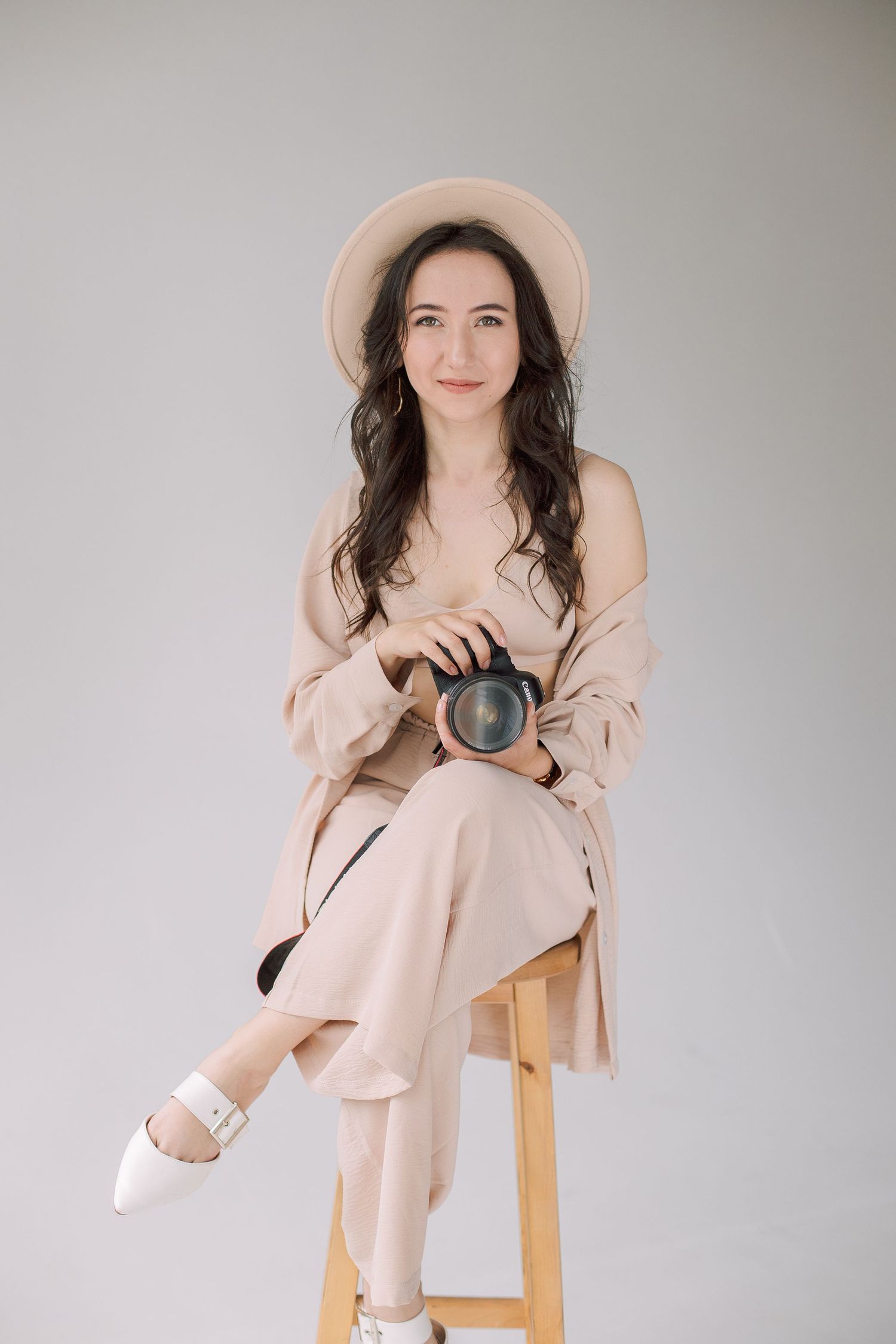 Photographer in pale pink outfit with hat sitting on wooden stool holding camera.