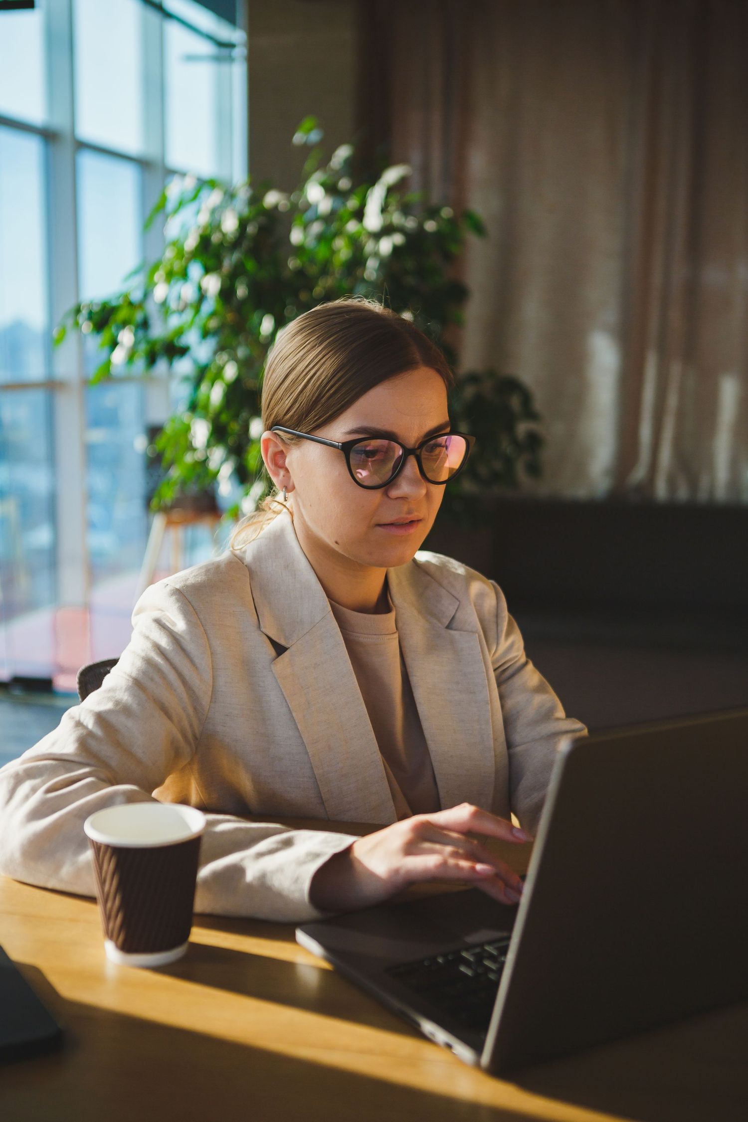 Professional working on laptop by window in business setting with coffee cup and indoor plants.