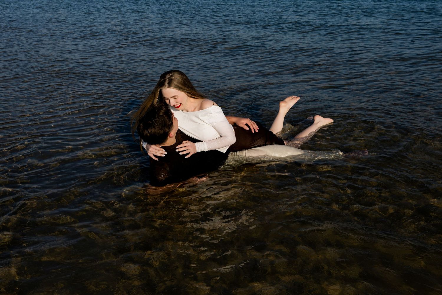 A figure reclines on dark rocks at the edge of a deep blue ocean during sunset.