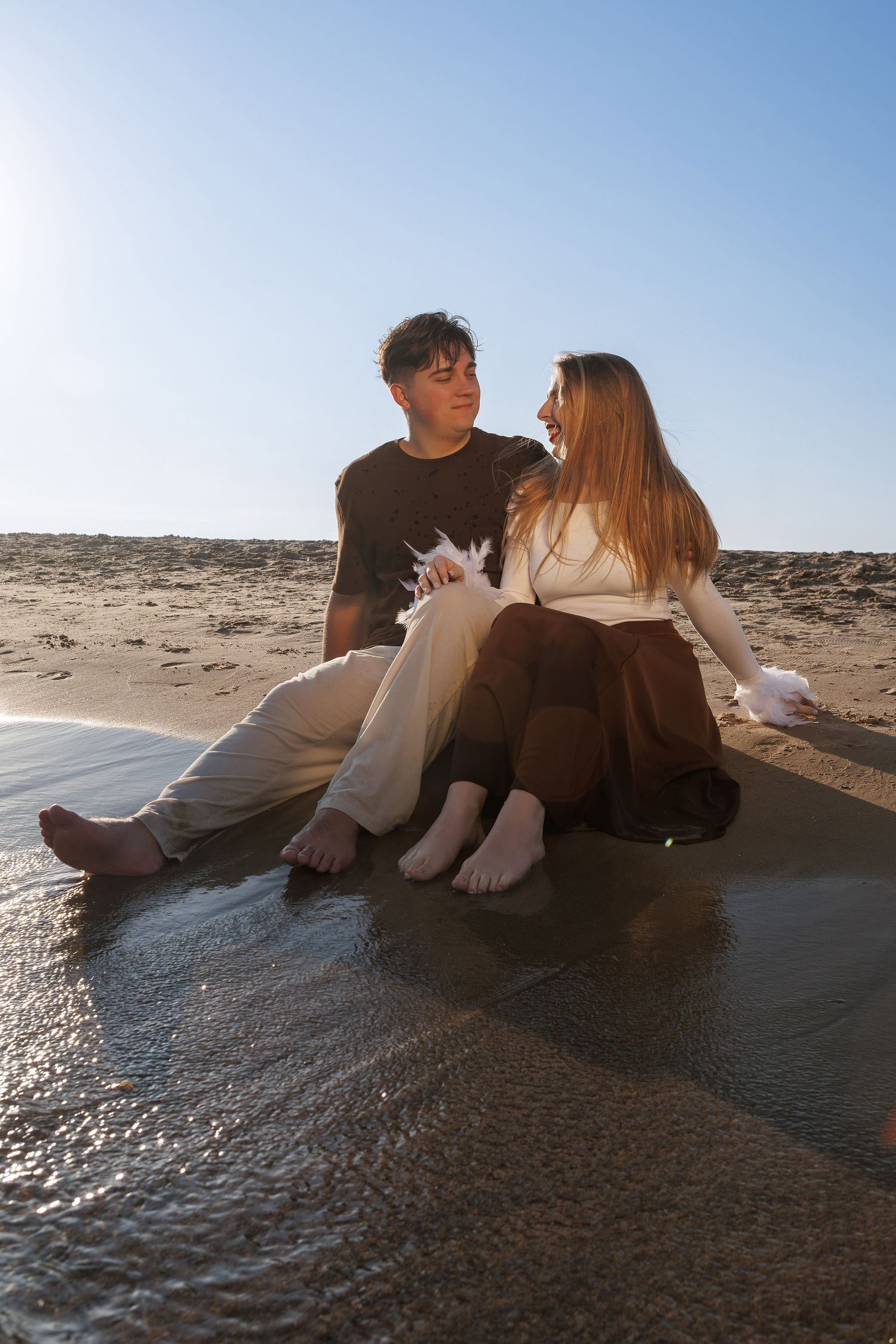 A couple sits together on a rocky beach during sunset creating a romantic silhouette against the blue sky.