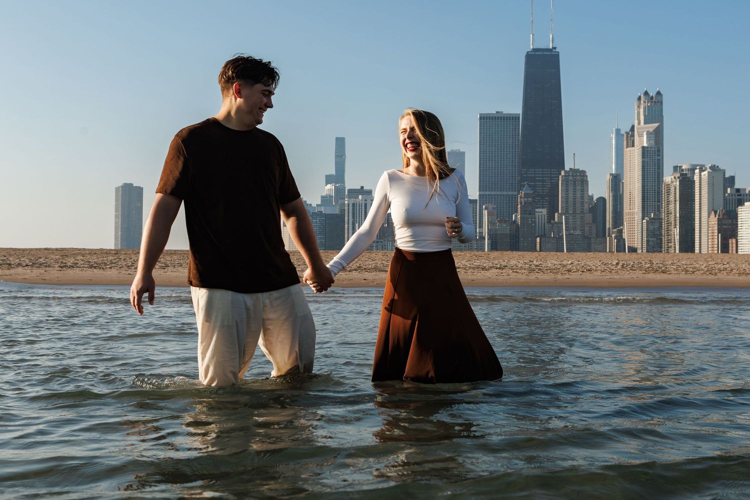 People wade through shallow water with a city skyline visible in the distance on a sunny day.