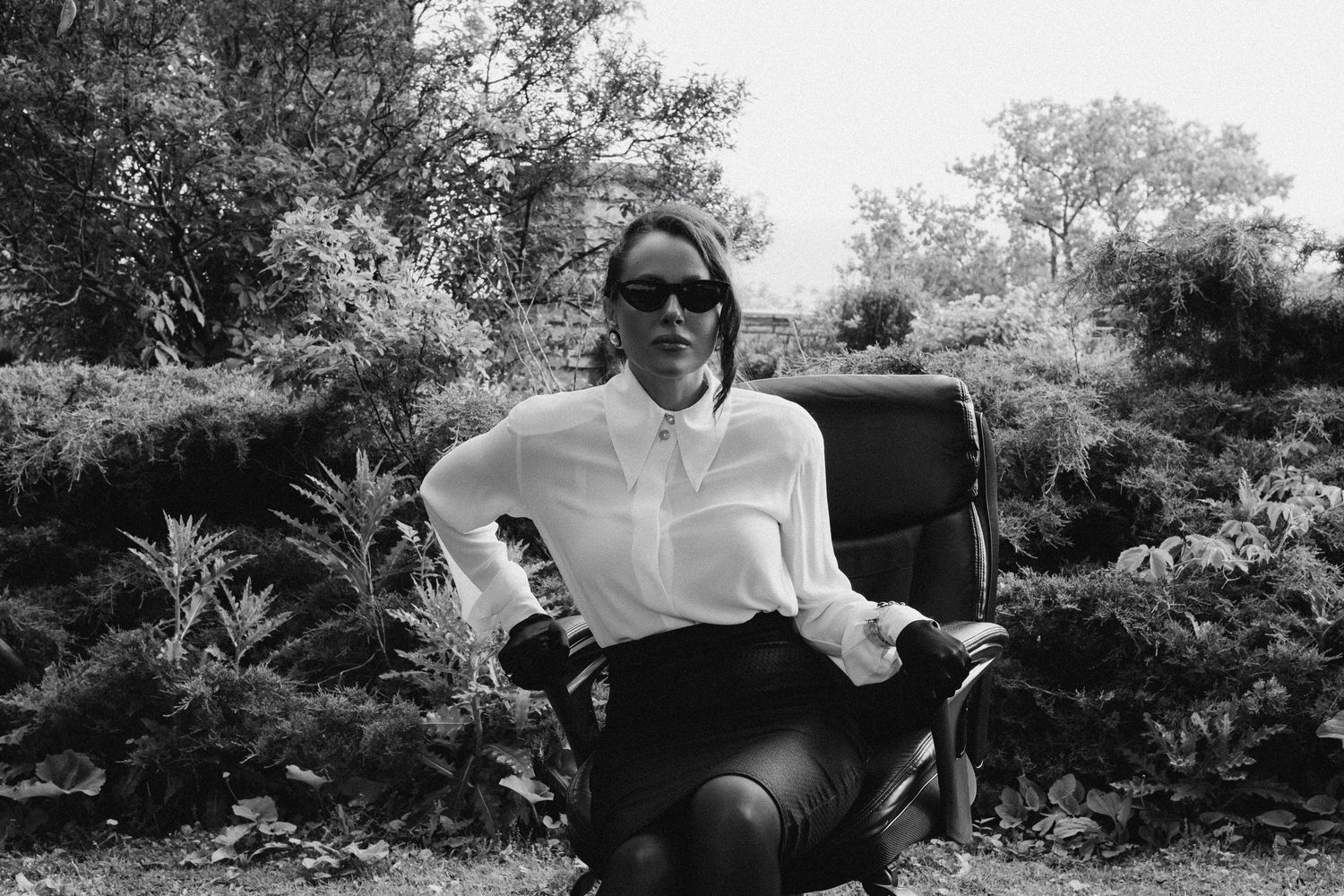 A person in sunglasses and white shirt sits relaxed in a chair outdoors surrounded by lush garden foliage.