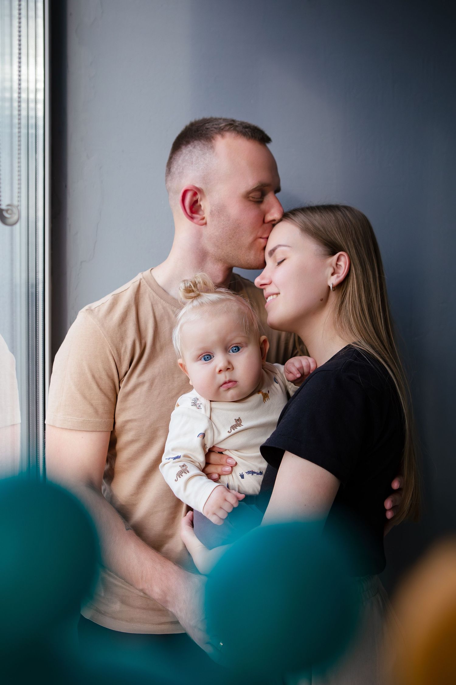 A tender family moment by a window as parents embrace while holding their baby in a beige and black color scheme.