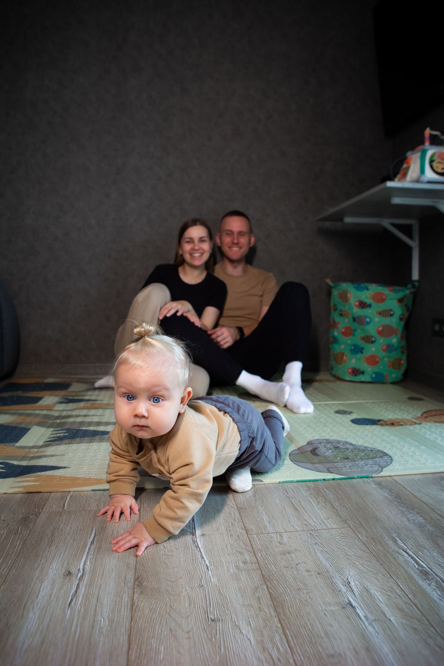 A baby in a tan onesie crawls on a gray wooden floor while adults sit in the background.