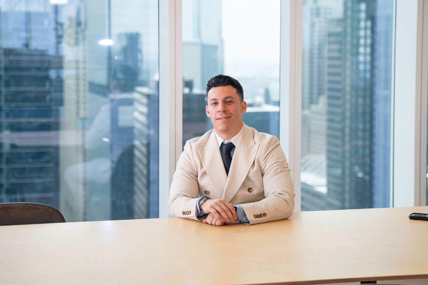 Professional in light suit sitting at conference table with city skyline view through windows.
