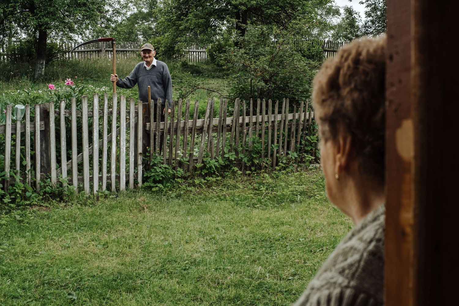 Person looking through doorway at someone standing by rustic wooden fence in rural garden.