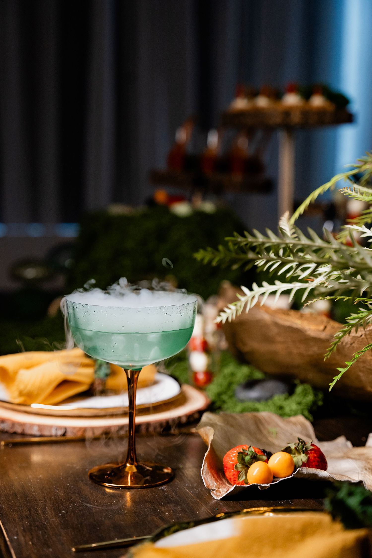 Elegant green cocktail with smoky effect served in copper-stemmed coupe glass among table decorations.