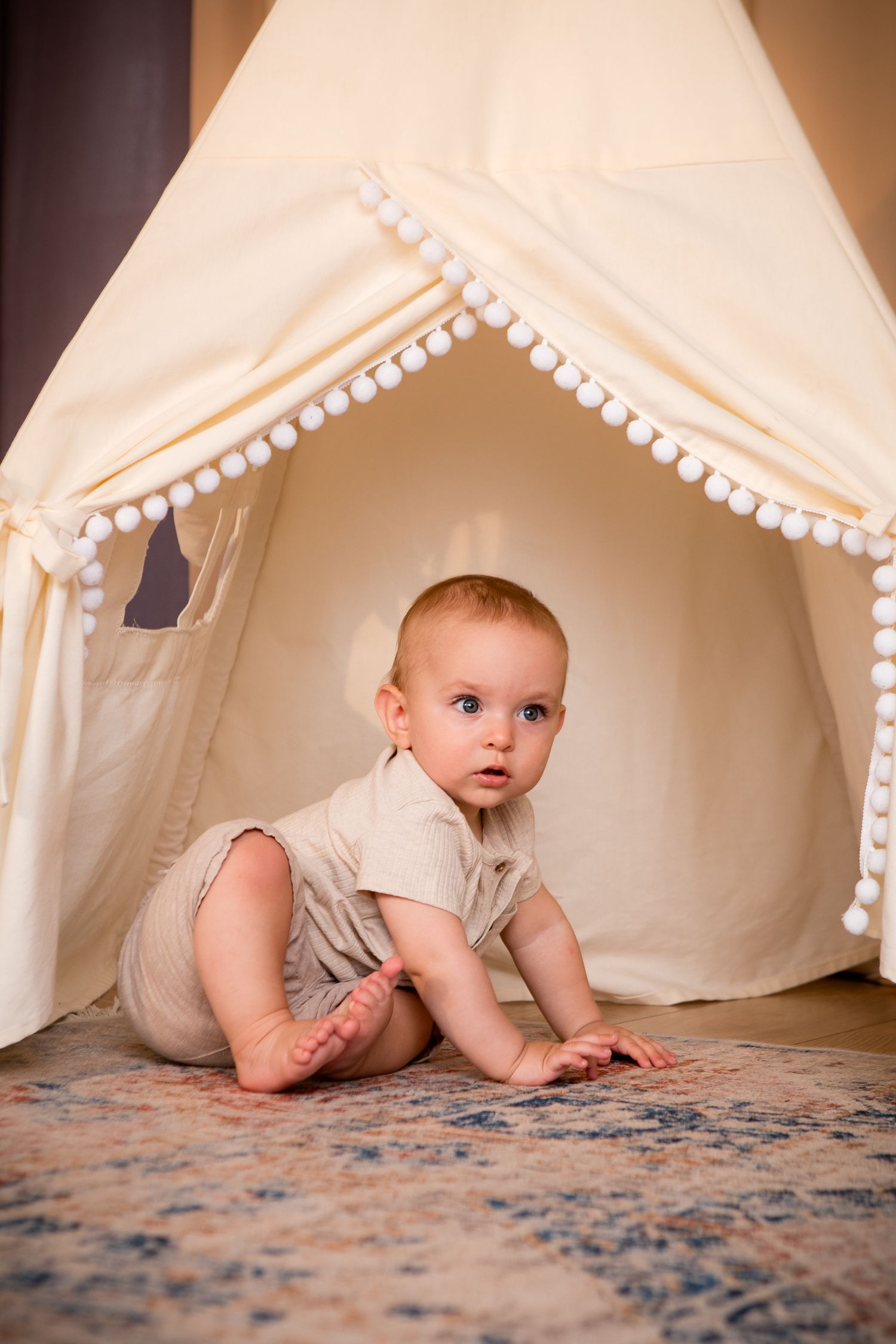 Baby crawling inside white teepee tent with pom pom trim.