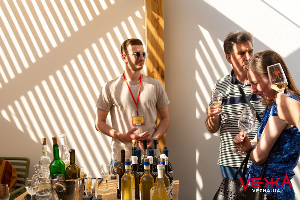 Two people taste wine in a sunlit room with dramatic shadow patterns on the wall.