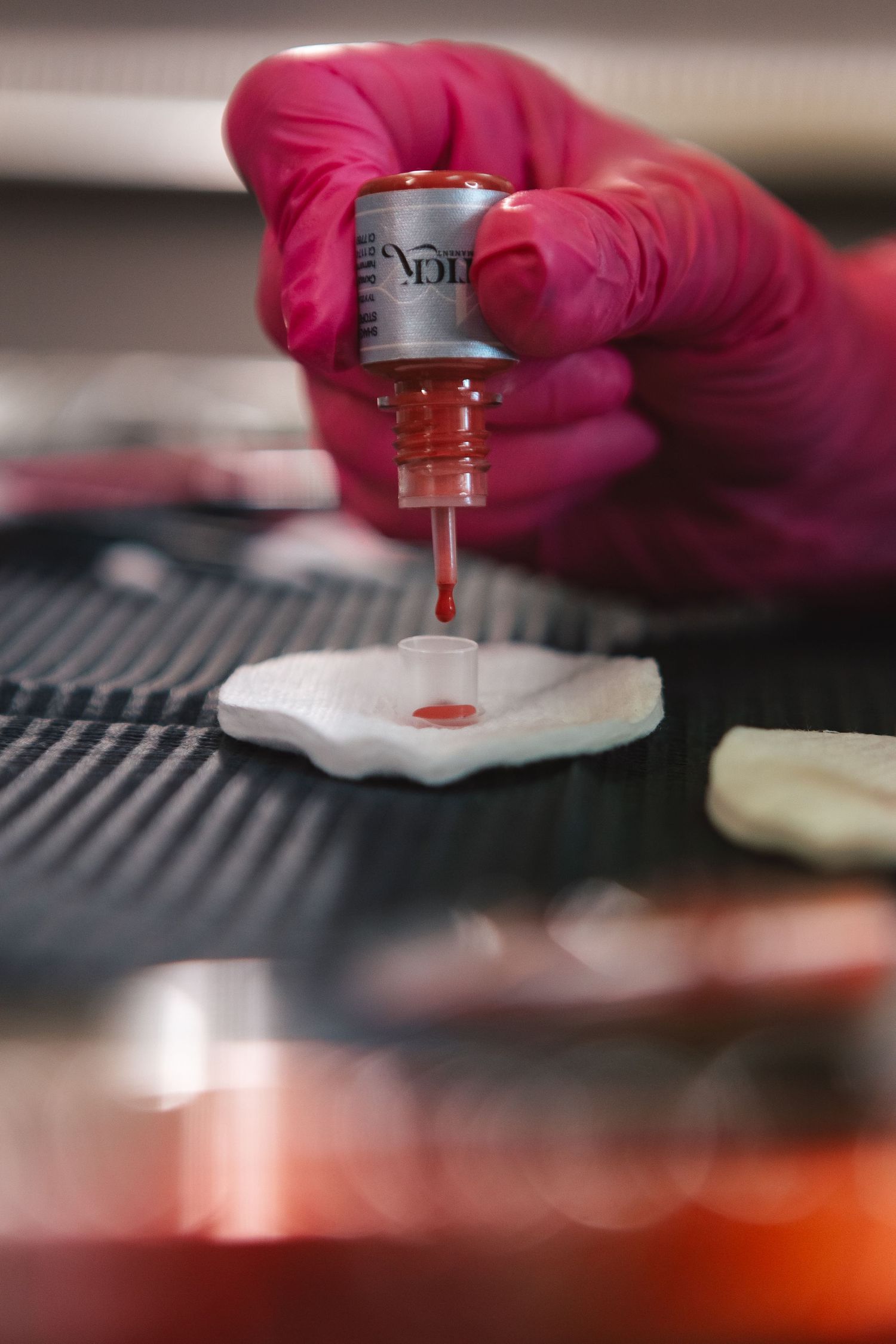 Pink-gloved hands dispensing red pigment from a small bottle onto a surface.