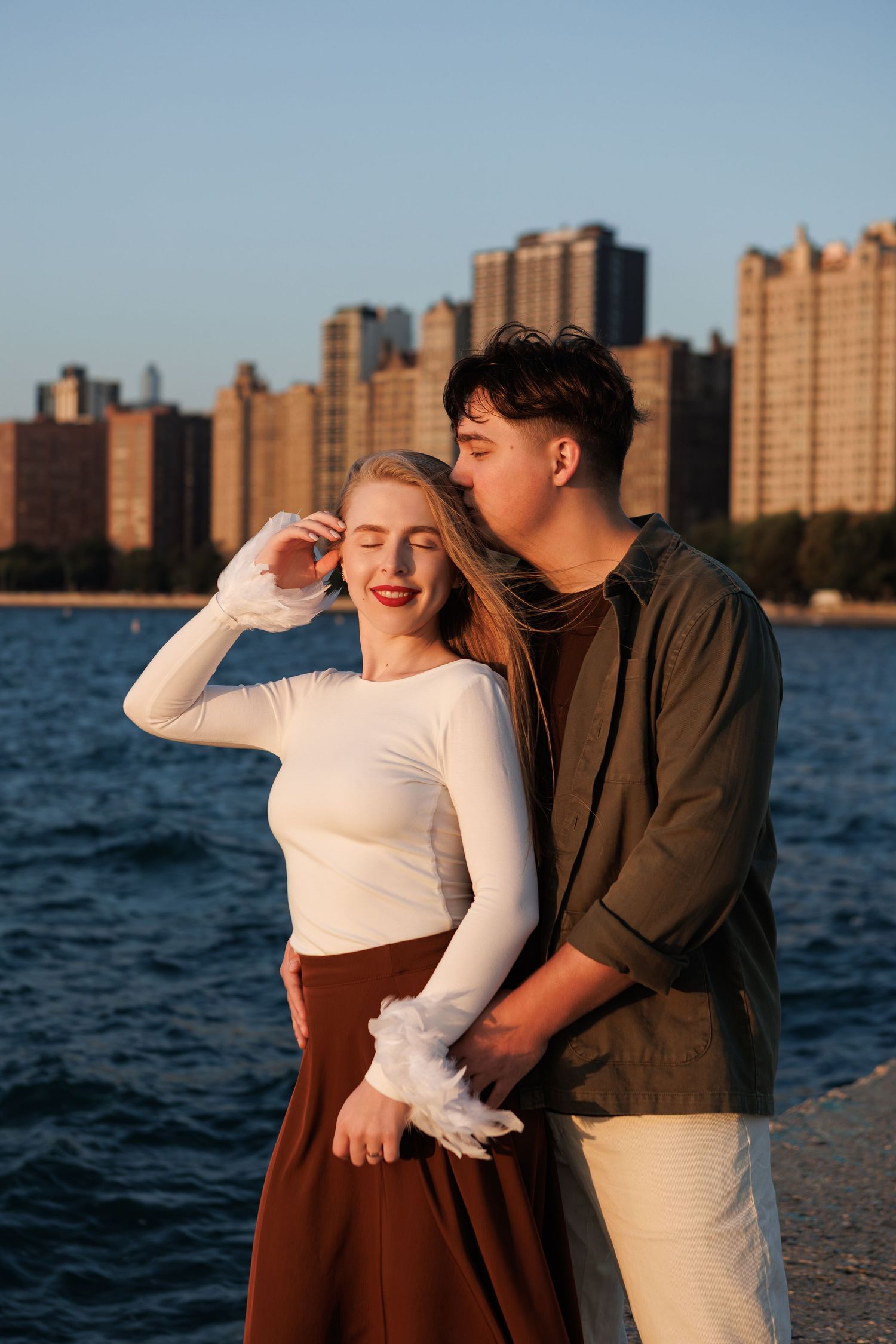 Couple sharing intimate moments by the waterfront with city skyline at sunset.