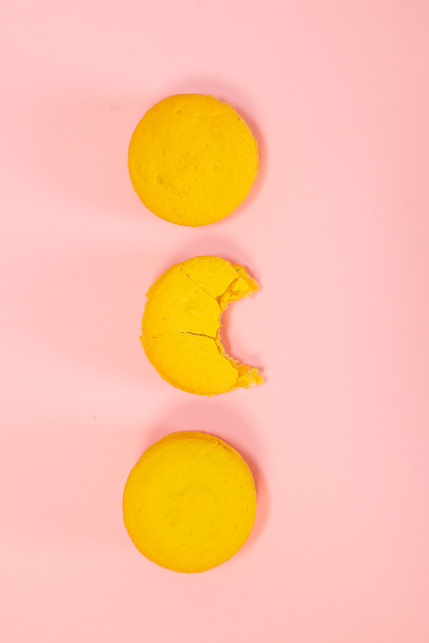 Three yellow macarons on pink background with one showing a bite mark.