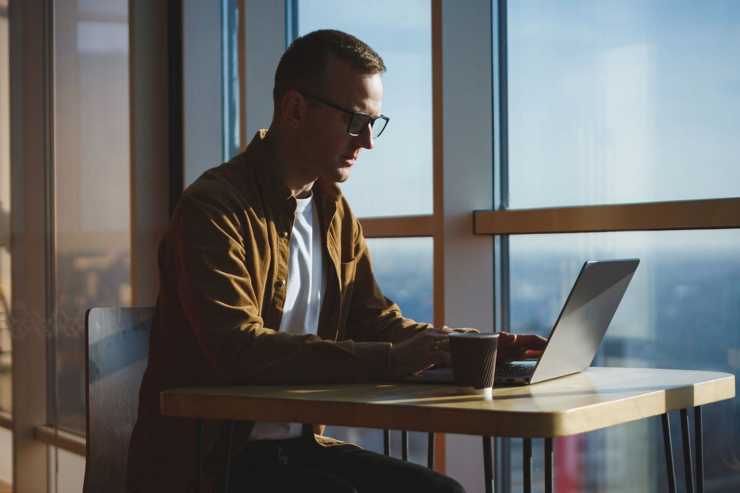 Person in casual attire working on laptop computer by large window during sunset.