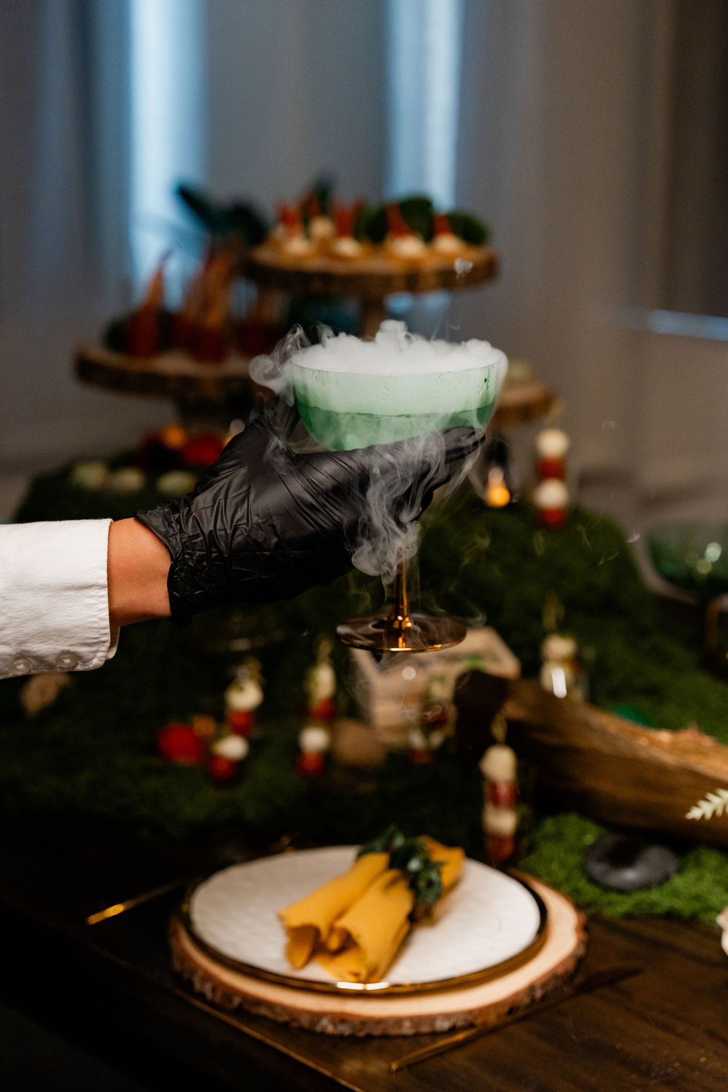 Black-gloved hand serving smoking cocktail at elegantly decorated table with greenery and candles.