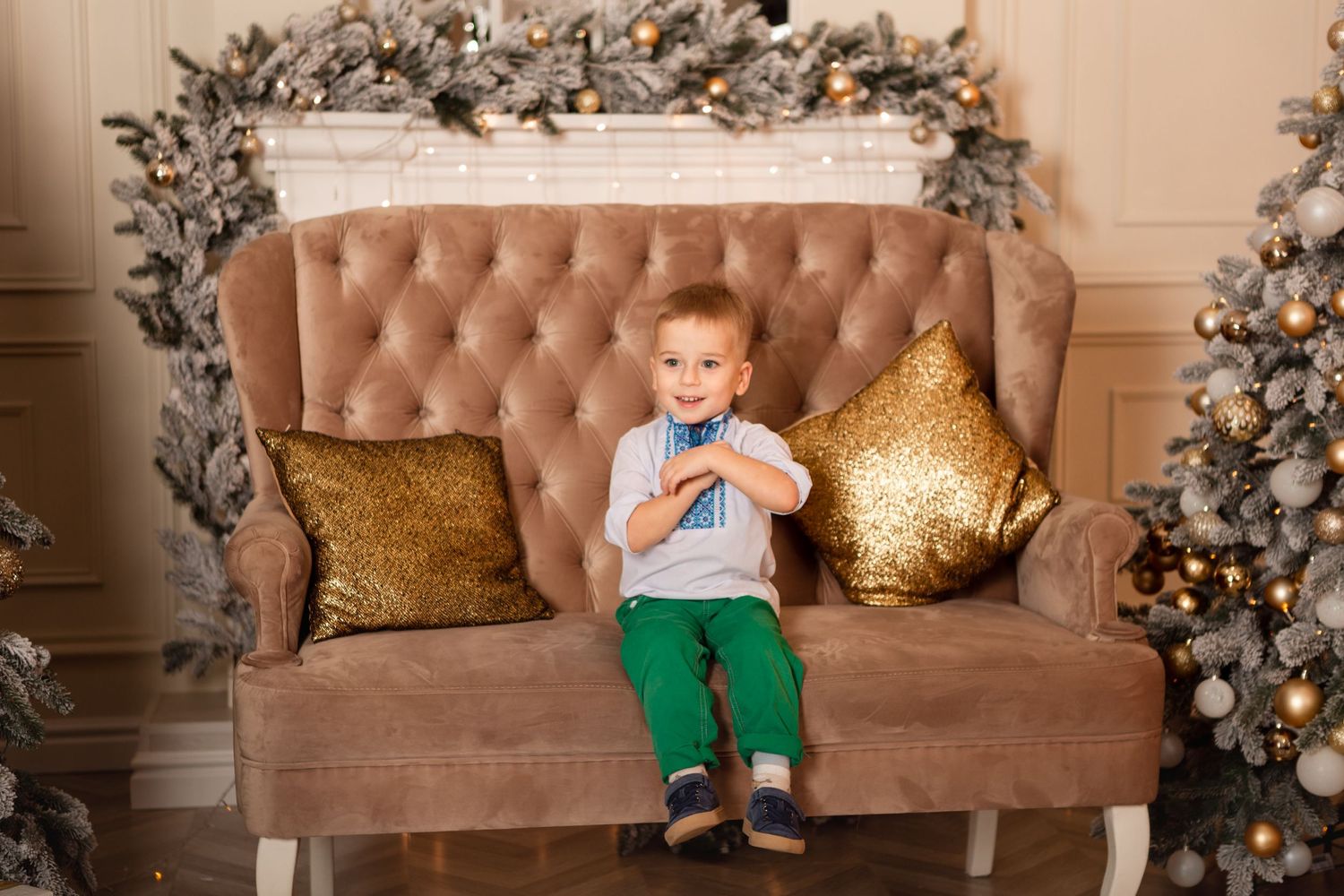 Young child sits on brown tufted sofa with gold pillows between decorated Christmas trees.