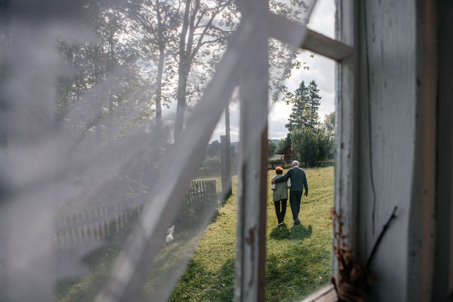Silhouette of a person seen through a curtained window in a sunlit garden.