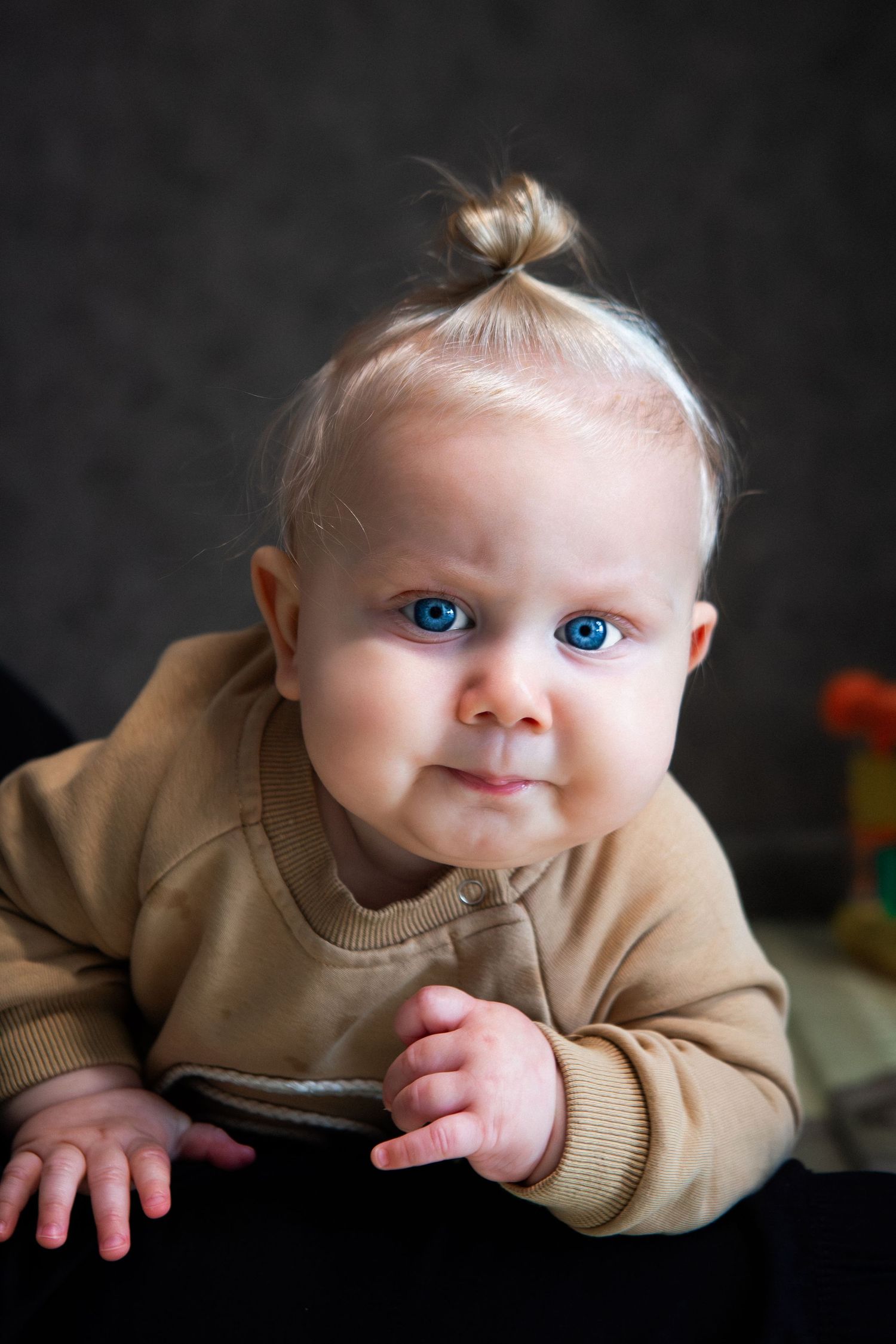 A baby in a tan sweater lies on their stomach against a dark background with striking blue eyes.