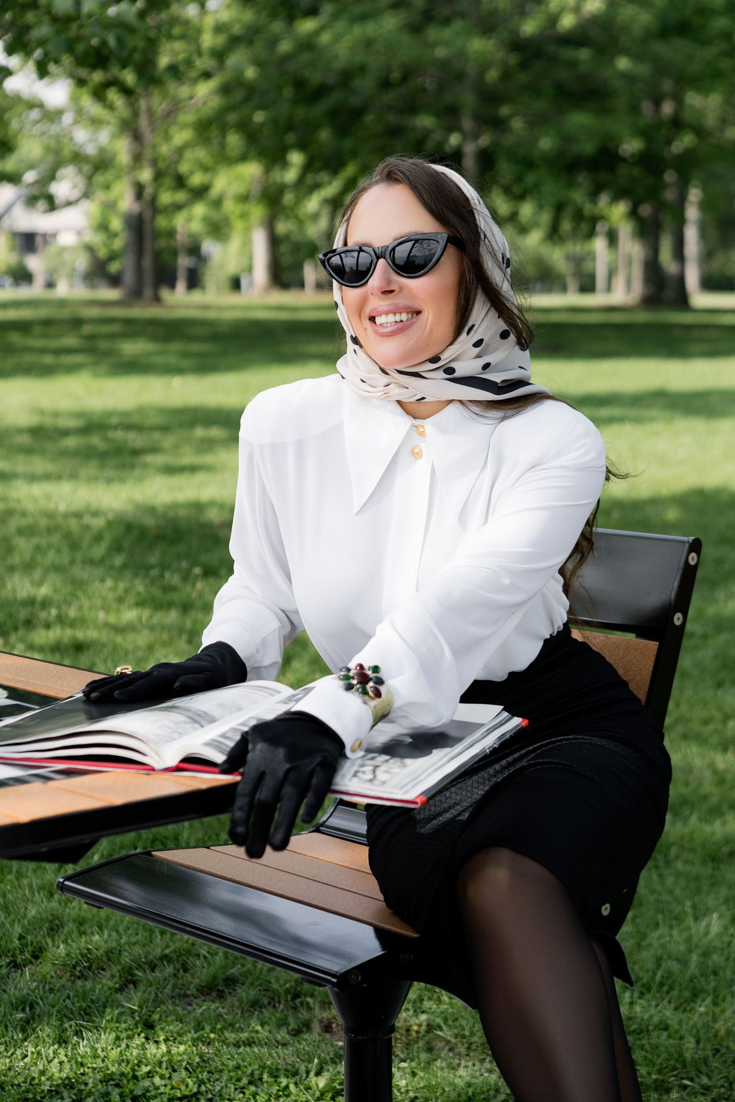 A person in a white shirt and dark tights lounges in a park with a book and sunglasses on a sunny day.